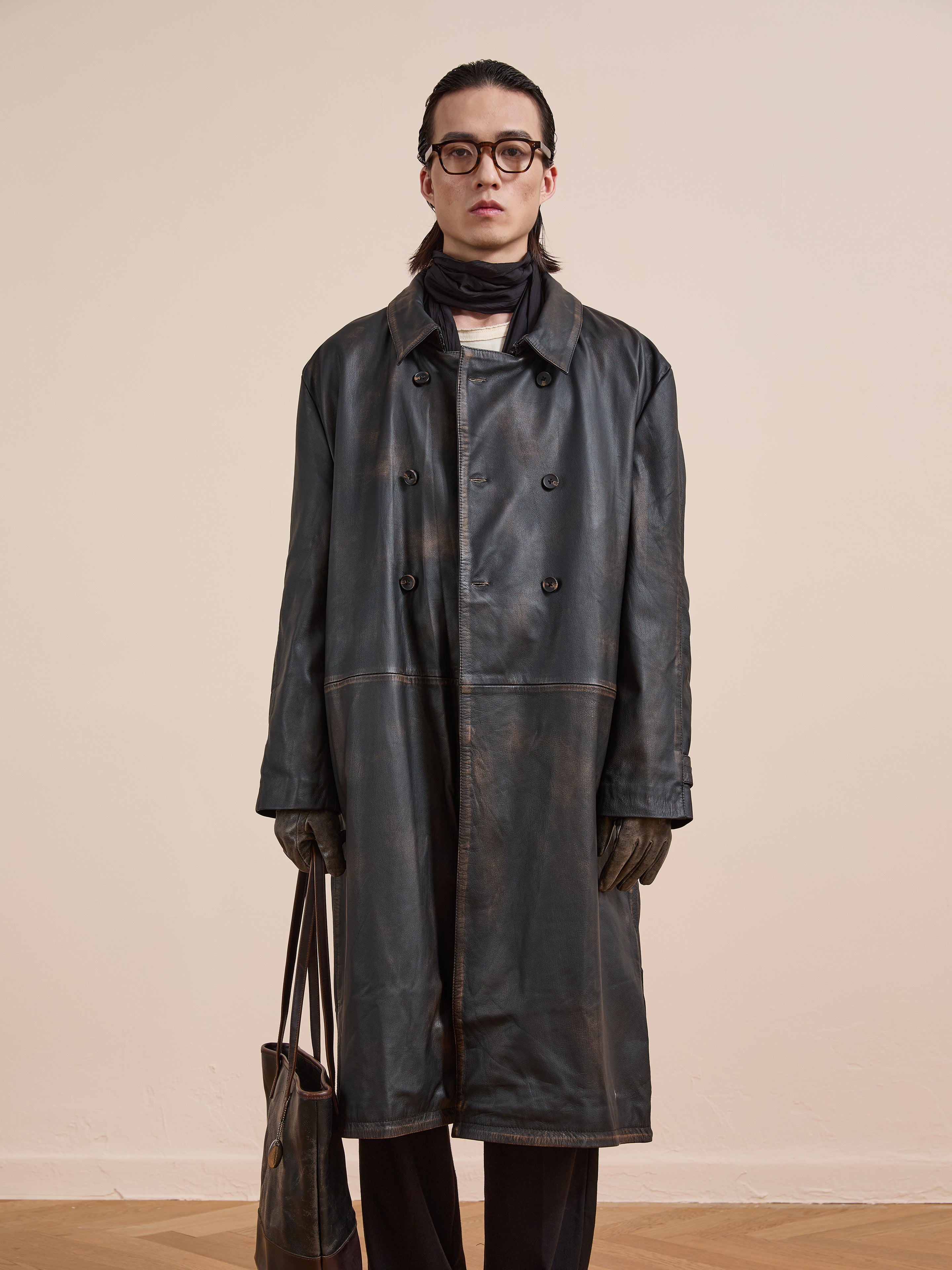A person stands indoors holding a brown bag, dressed in glasses, gloves, a scarf, and the FOUND Distressed Leather Trench Coat.