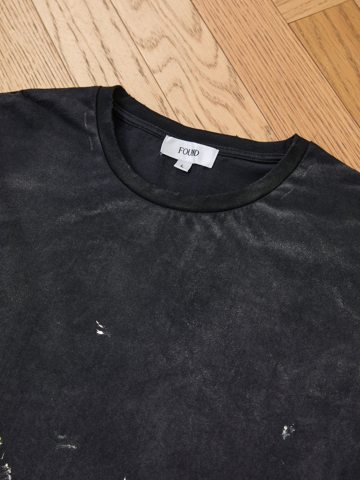 A black FOUND Distressed Paint Tee rests on a wooden floor. This unisex shirt features a round neckline, subtle paint details and wear marks, and is designed with a relaxed fit.