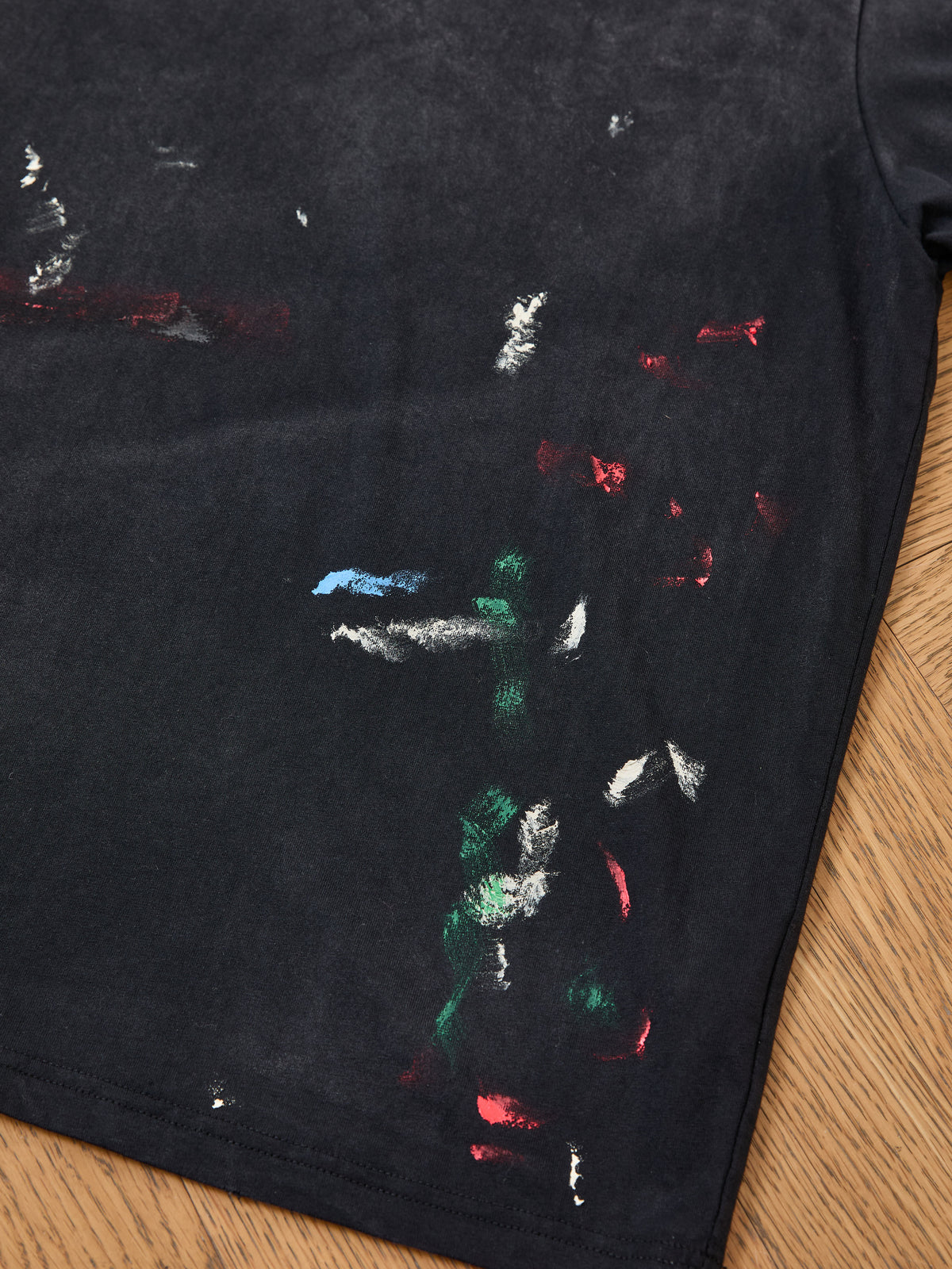 FOUND Distressed Paint Tee in black, featuring a unisex relaxed fit and scattered red, green, blue, and white paint smudges on the lower right side. Photographed on a wooden surface, this tee offers a distressed, lived-in look ideal for casual styling.