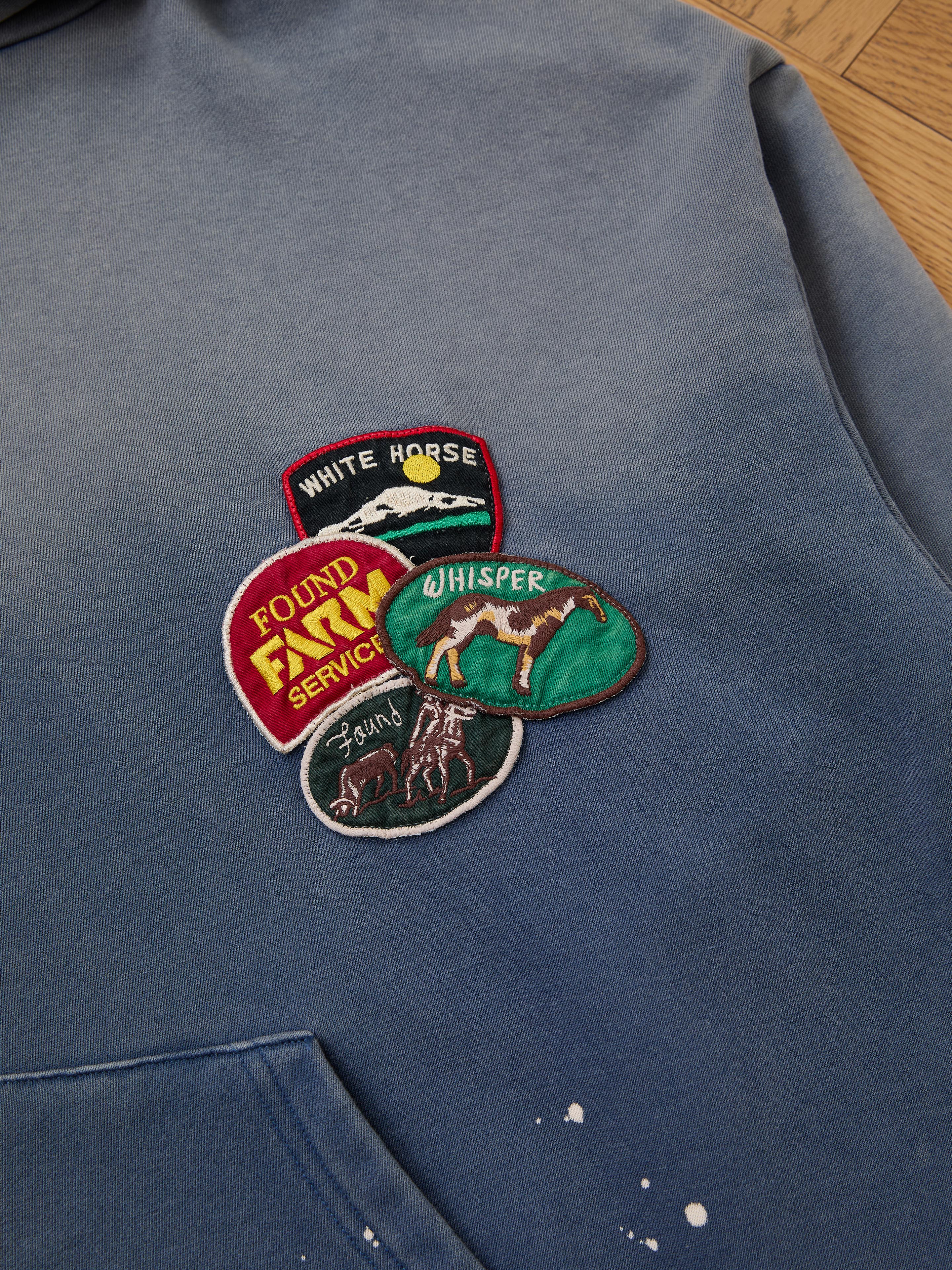 The FOUND Distressed Patch Hoodie is a vintage-style blue French Terry cotton hoodie featuring four colorful embroidered patches: a white horse, "Found Fun Service," another horse, and a sun.