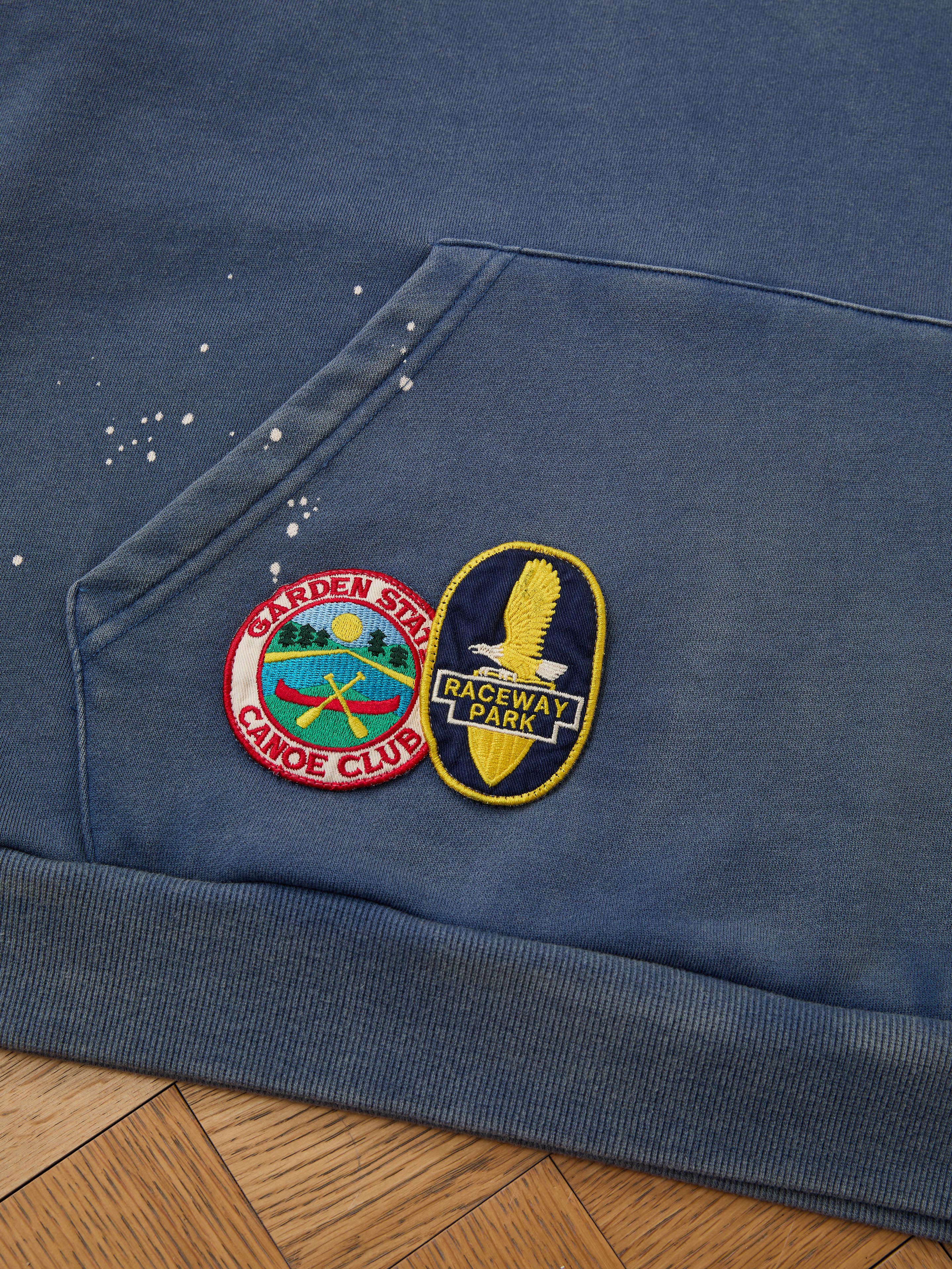 The FOUND Distressed Patch Hoodie in blue features two embroidered patches: one with "Garden State Canoe Club" and crossed paddles, and another reading "Raceway Park" with a winged tire.