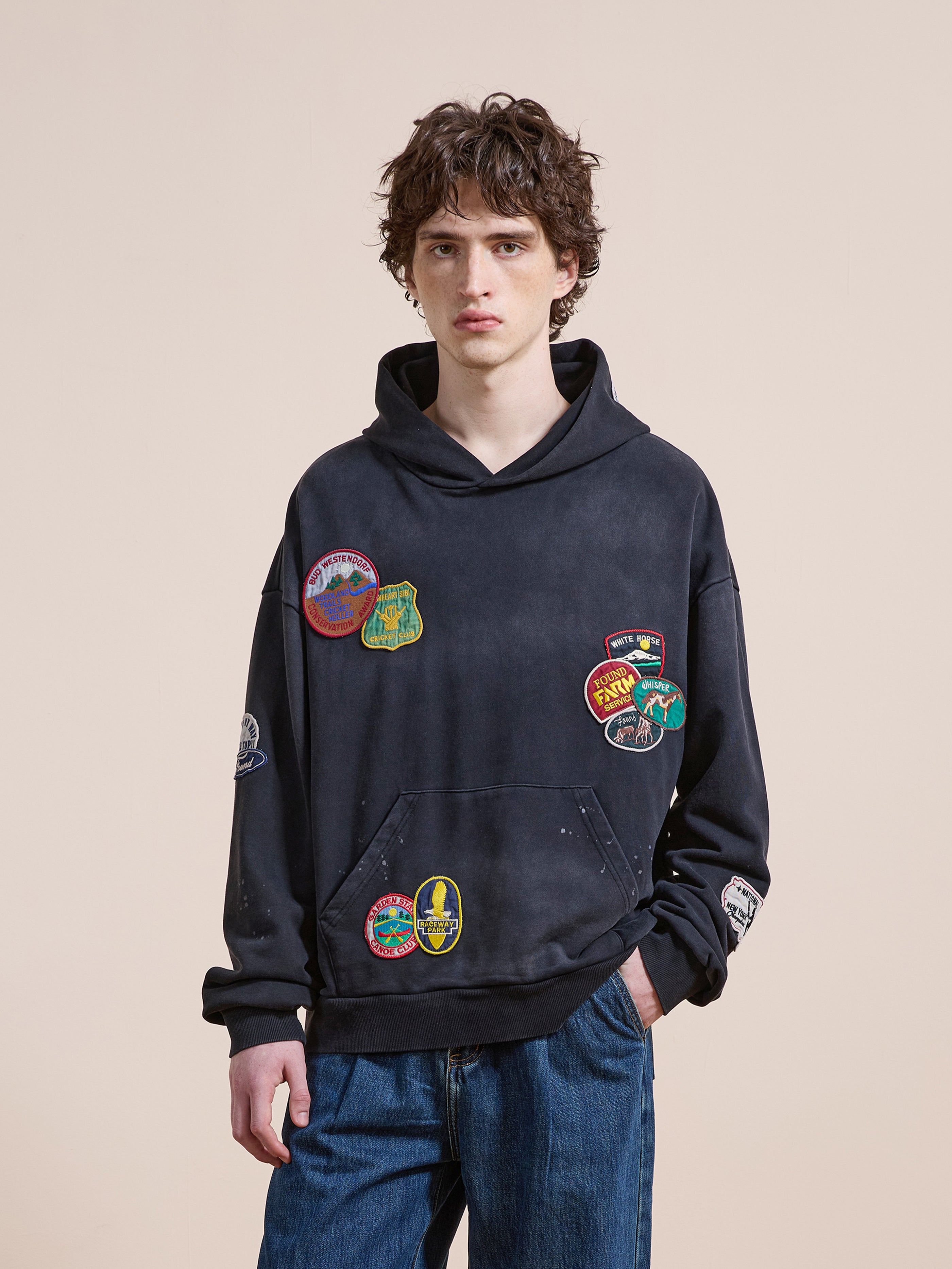 A young person in the FOUND Distressed Patch Hoodie—a unisex relaxed fit with colorful embroidered patches—paired with blue jeans stands against a beige background, gazing at the camera.