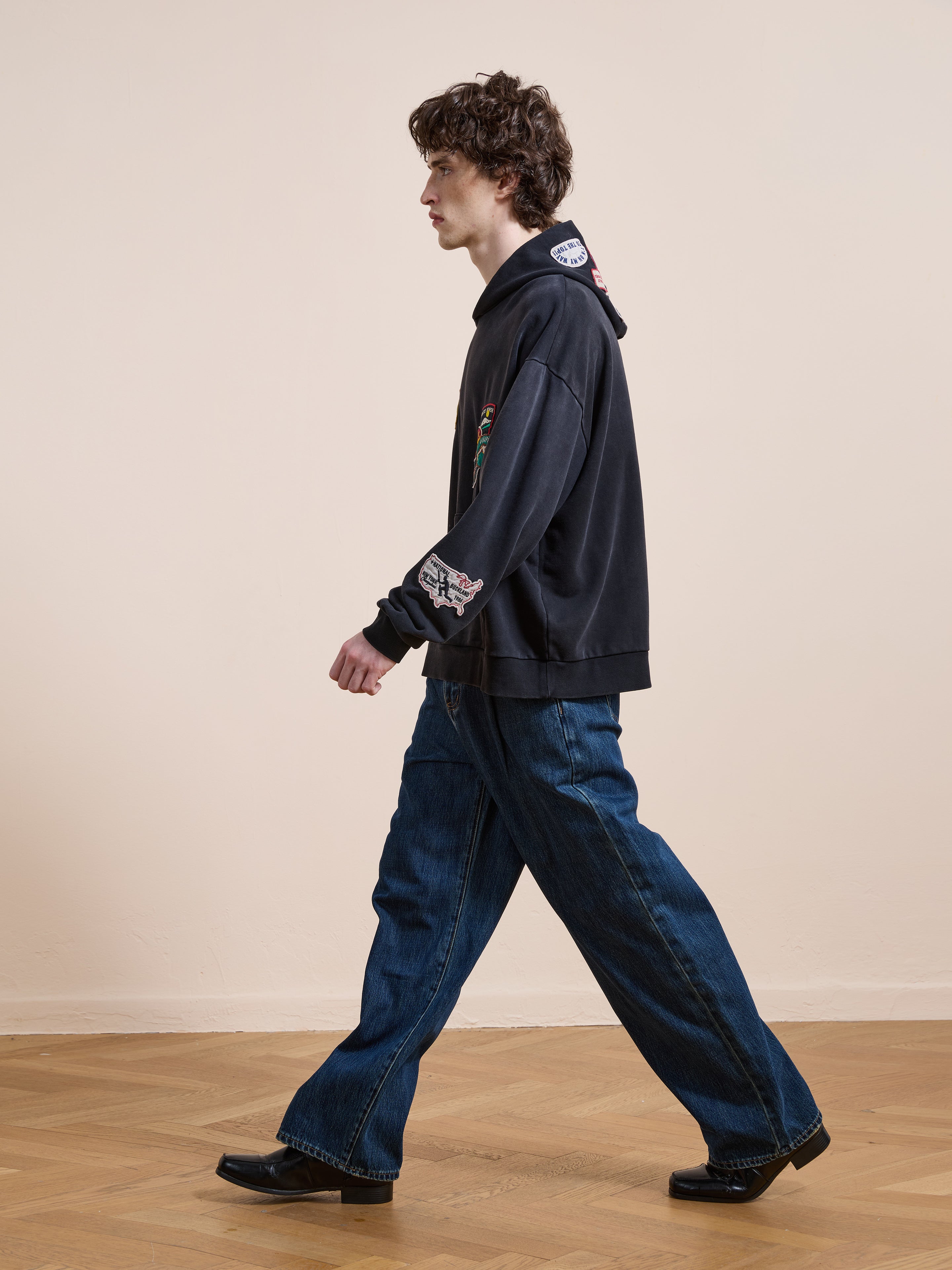 A person with curly hair walks indoors on a wooden floor, wearing the FOUND Distressed Patch Hoodie in vintage washed black, paired with loose blue jeans and black shoes against a plain light wall.