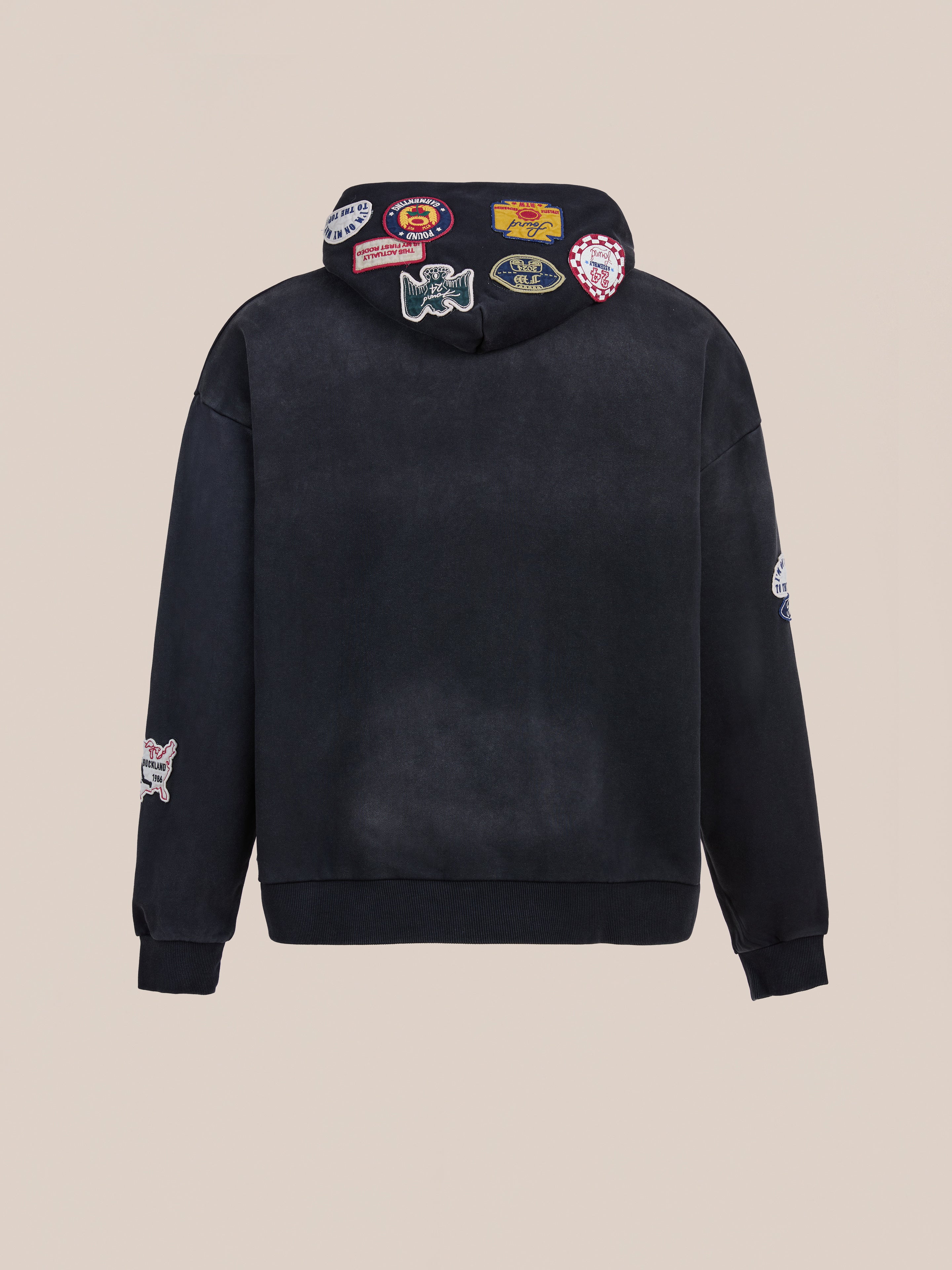 The FOUND Distressed Patch Hoodie is a unisex relaxed fit black sweatshirt shown from the back, with colorful embroidered patches on the hood and sleeves, set against a beige background.