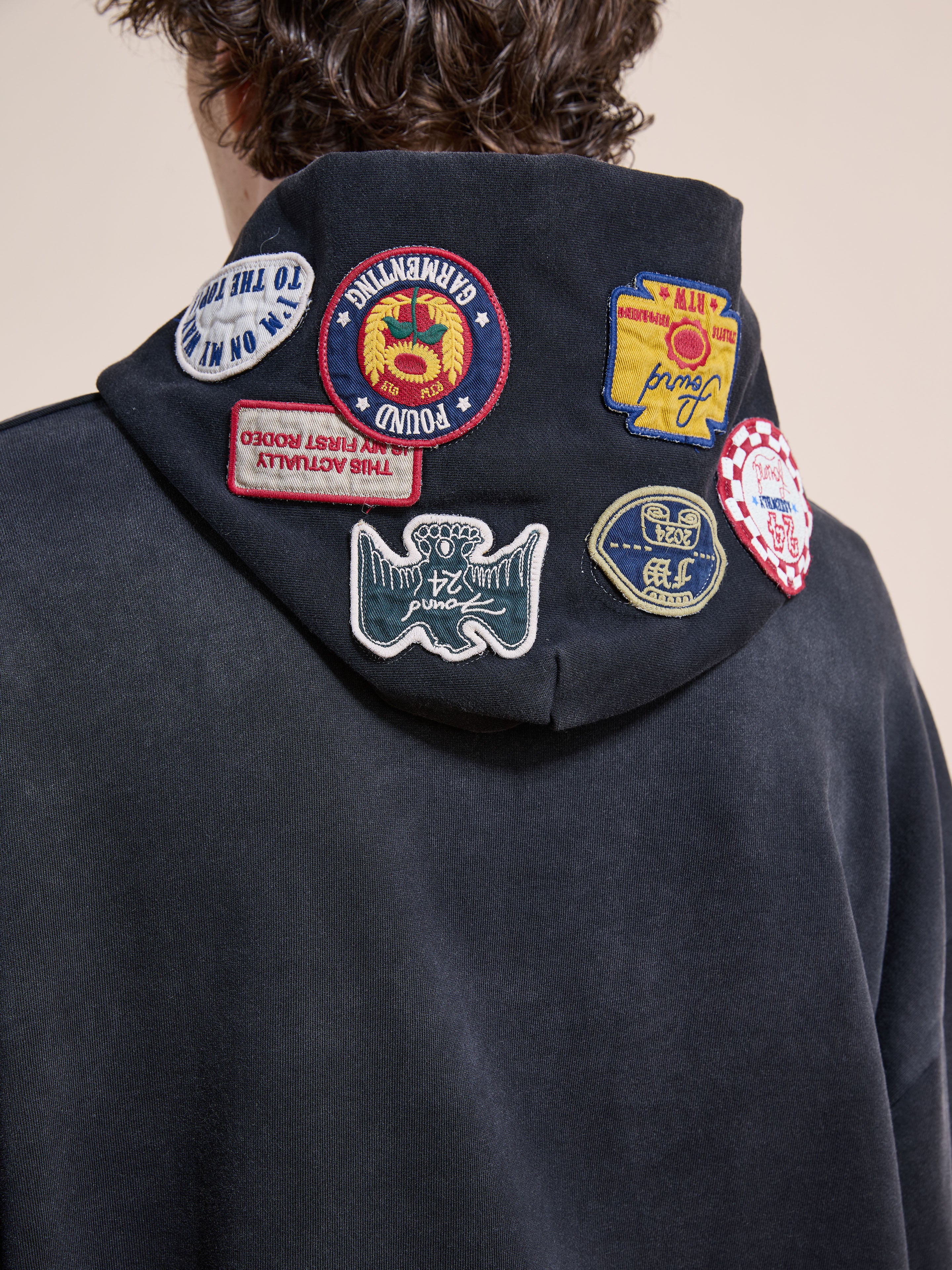 A person is seen from behind wearing the FOUND Distressed Patch Hoodie, a unisex relaxed-fit vintage washed black hoodie featuring multiple colorful embroidered patches on the hood.