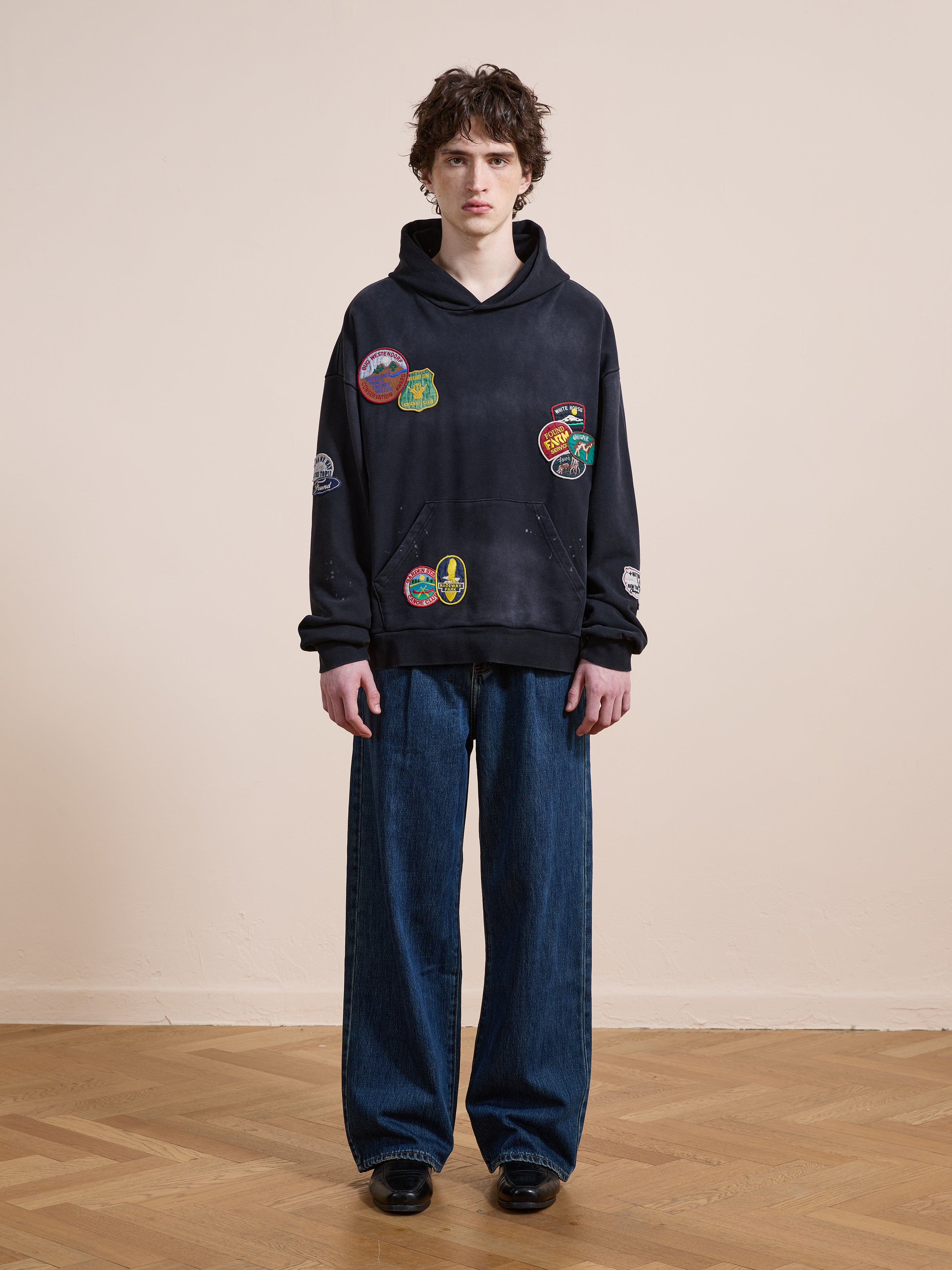 A person stands indoors wearing FOUND's Distressed Patch Hoodie, a unisex relaxed fit in vintage washed black with embroidered patches, paired with wide-leg blue jeans for an effortless, modern look.