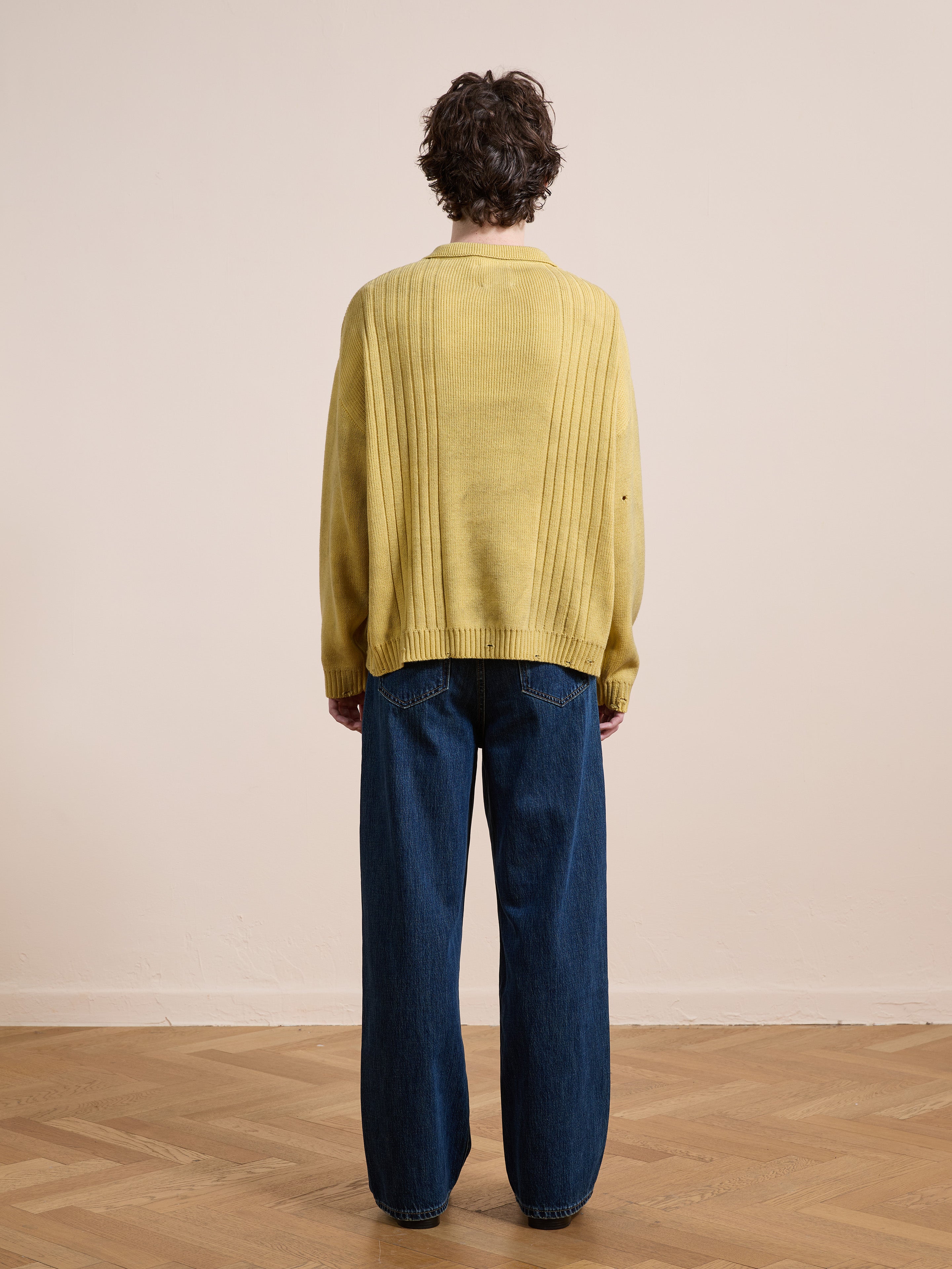 A person stands indoors facing away from the camera, wearing the FOUND Distressed Pullover Knit sweater and wide-leg blue jeans, on a wooden floor against a plain light-colored wall.