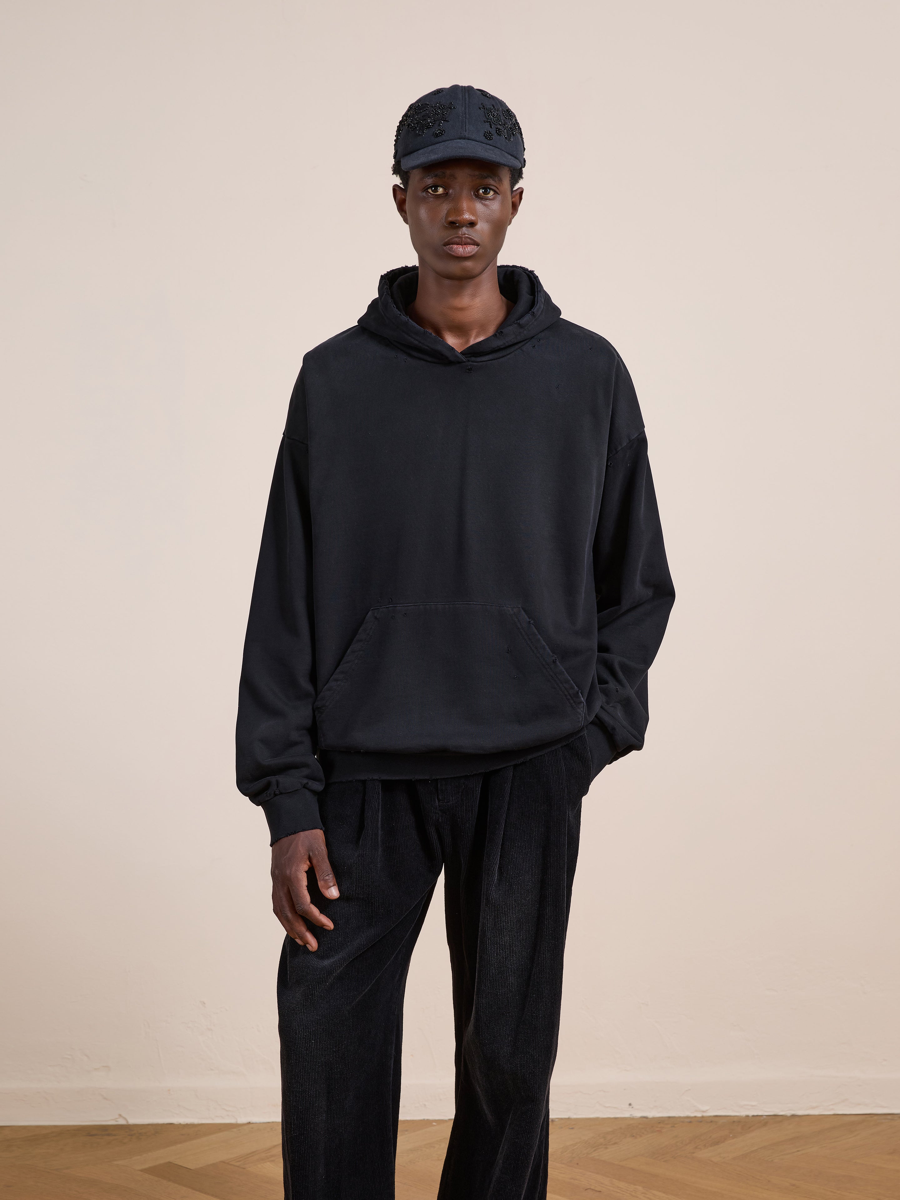 A person wears the FOUND Distressed Pullover Hoodie in black, paired with black pants and a black cap, standing indoors against a plain light-colored wall.