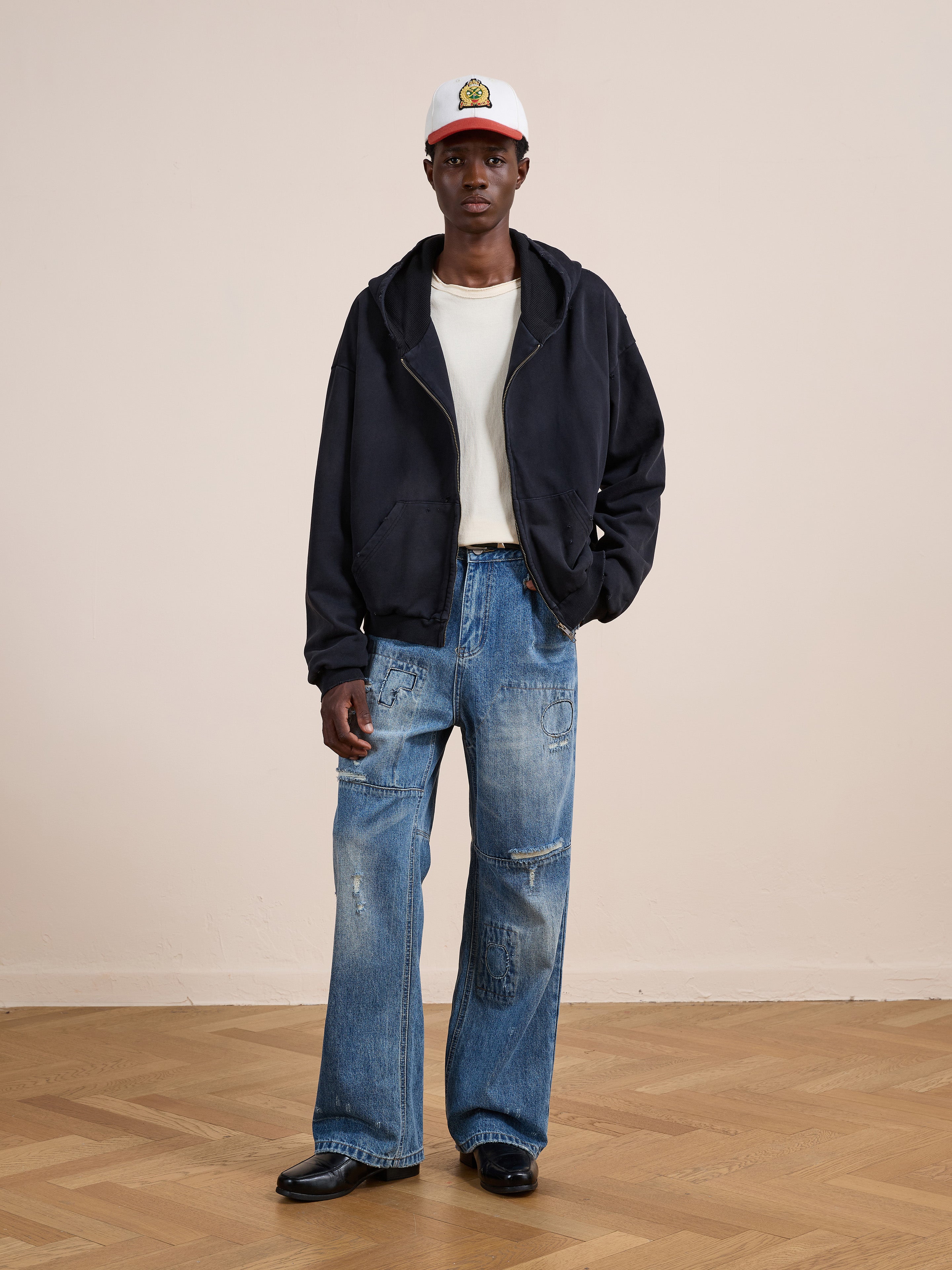 A person stands indoors wearing the FOUND Distressed Thermal Lined Zip Up Hoodie, paired with a white cap, white t-shirt, loose ripped blue jeans, and black shoes on a wooden floor against a plain light-colored wall.