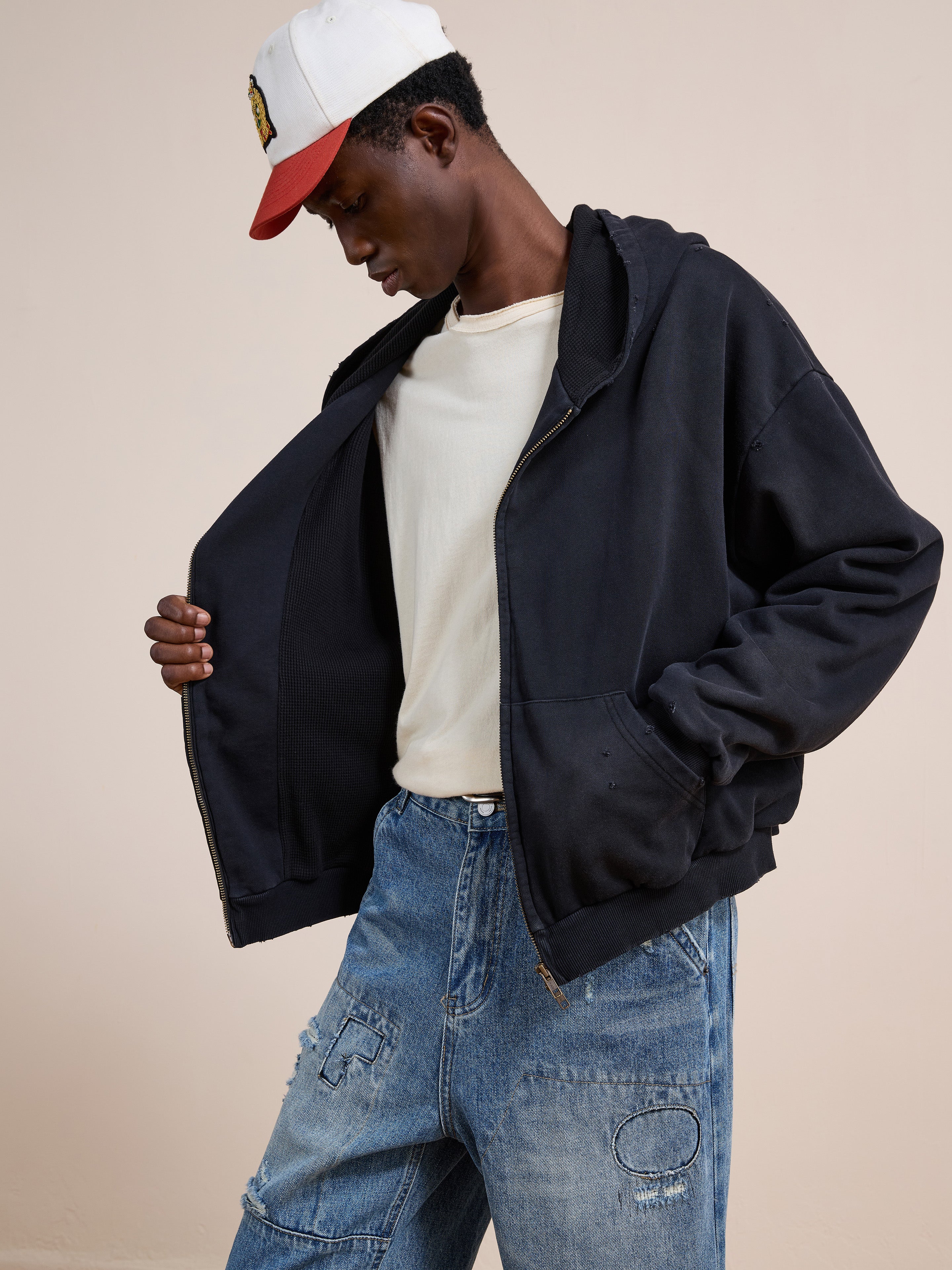 A person wearing a white cap, white t-shirt, FOUND Distressed Thermal Lined Zip Up Hoodie, and baggy blue jeans looks down while holding open the sides of their hoodie.