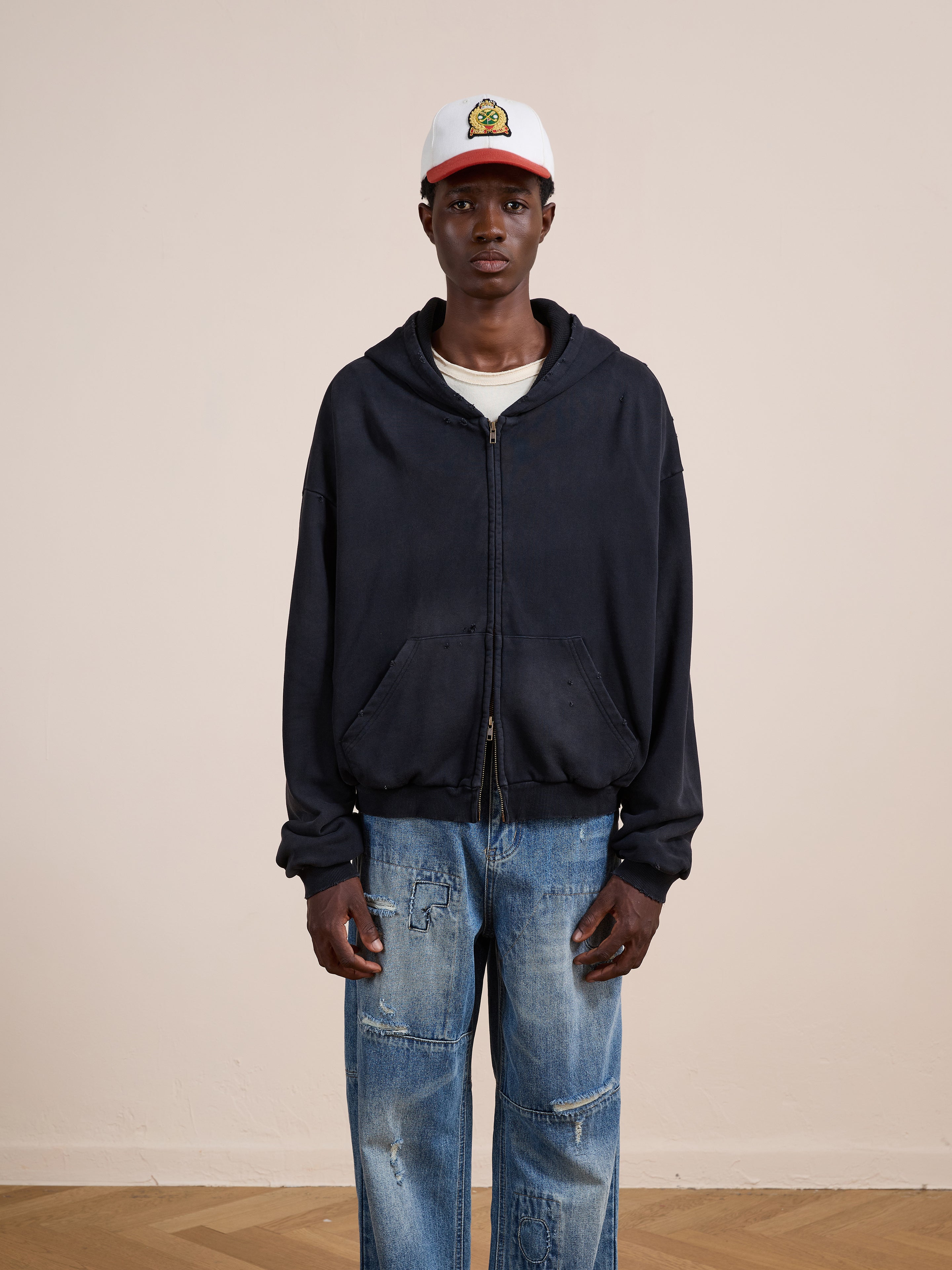 A person in the FOUND Distressed Thermal Lined Zip Up Hoodie, layered over a white shirt, paired with a white cap and blue distressed jeans, stands against a plain light-colored wall.