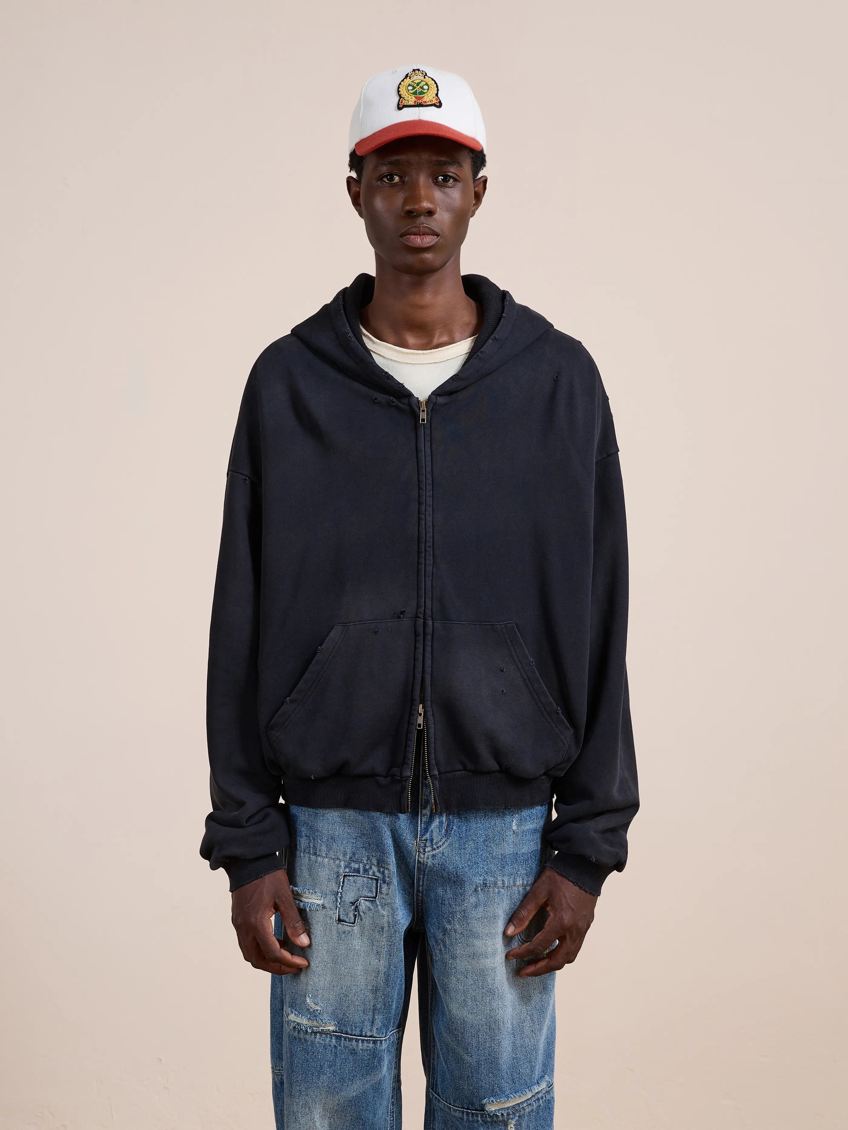 A person wears the FOUND Distressed Thermal Lined Zip Up Hoodie in washed black, a white cap, white t-shirt, and blue jeans while standing against a plain beige background.