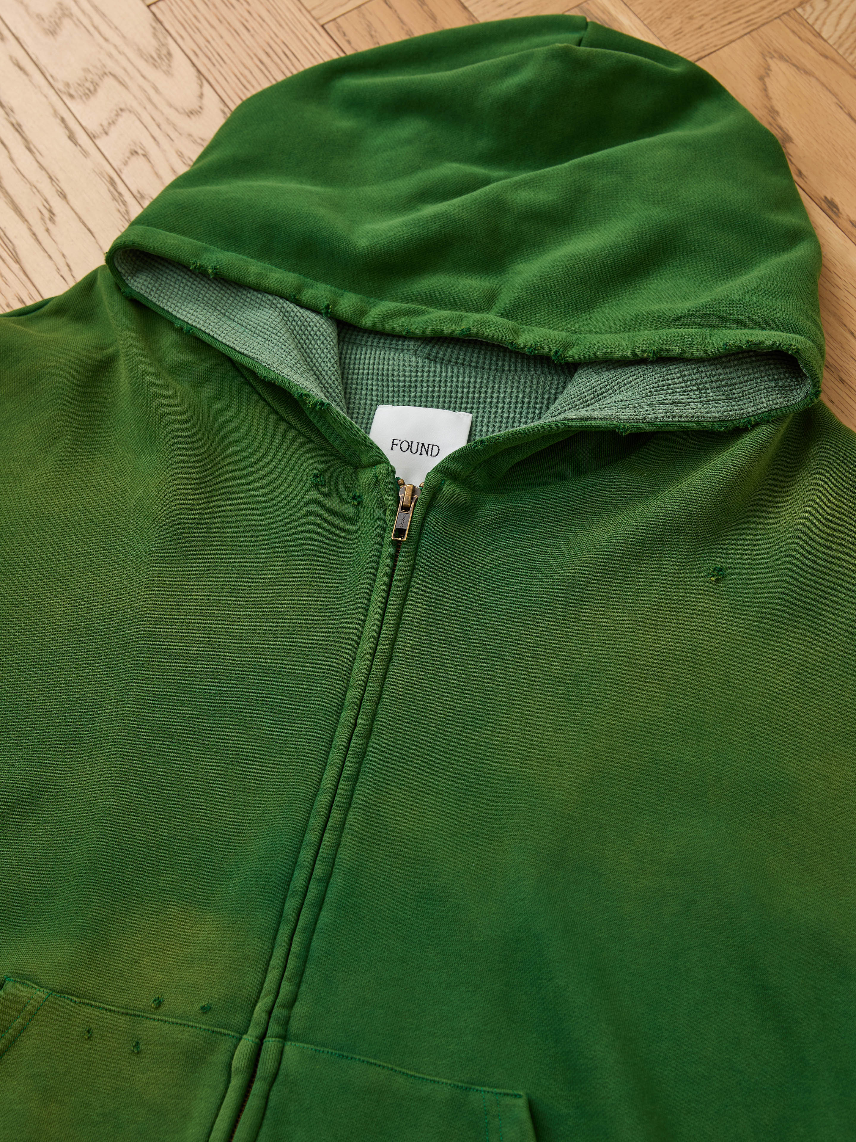The FOUND Distressed Thermal Lined Zip Up Hoodie in green features a cozy waffle-knit interior, thermal-lined hood, minor distressing, and a relaxed unisex fit. Shown laid flat on wood to highlight its stylish comfort.