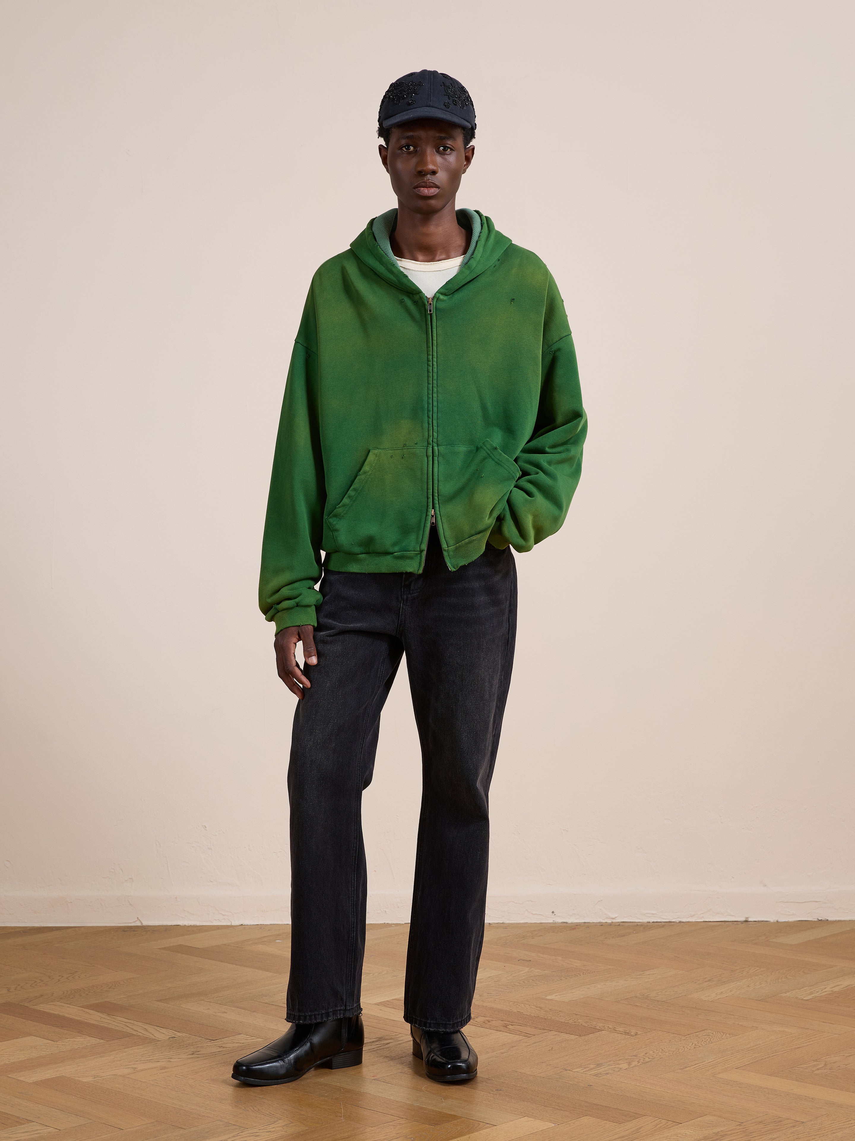 A person in a green FOUND Distressed Thermal Lined Zip Up Hoodie, black jeans, black shoes, and a navy cap stands on a wooden floor against a plain beige wall.