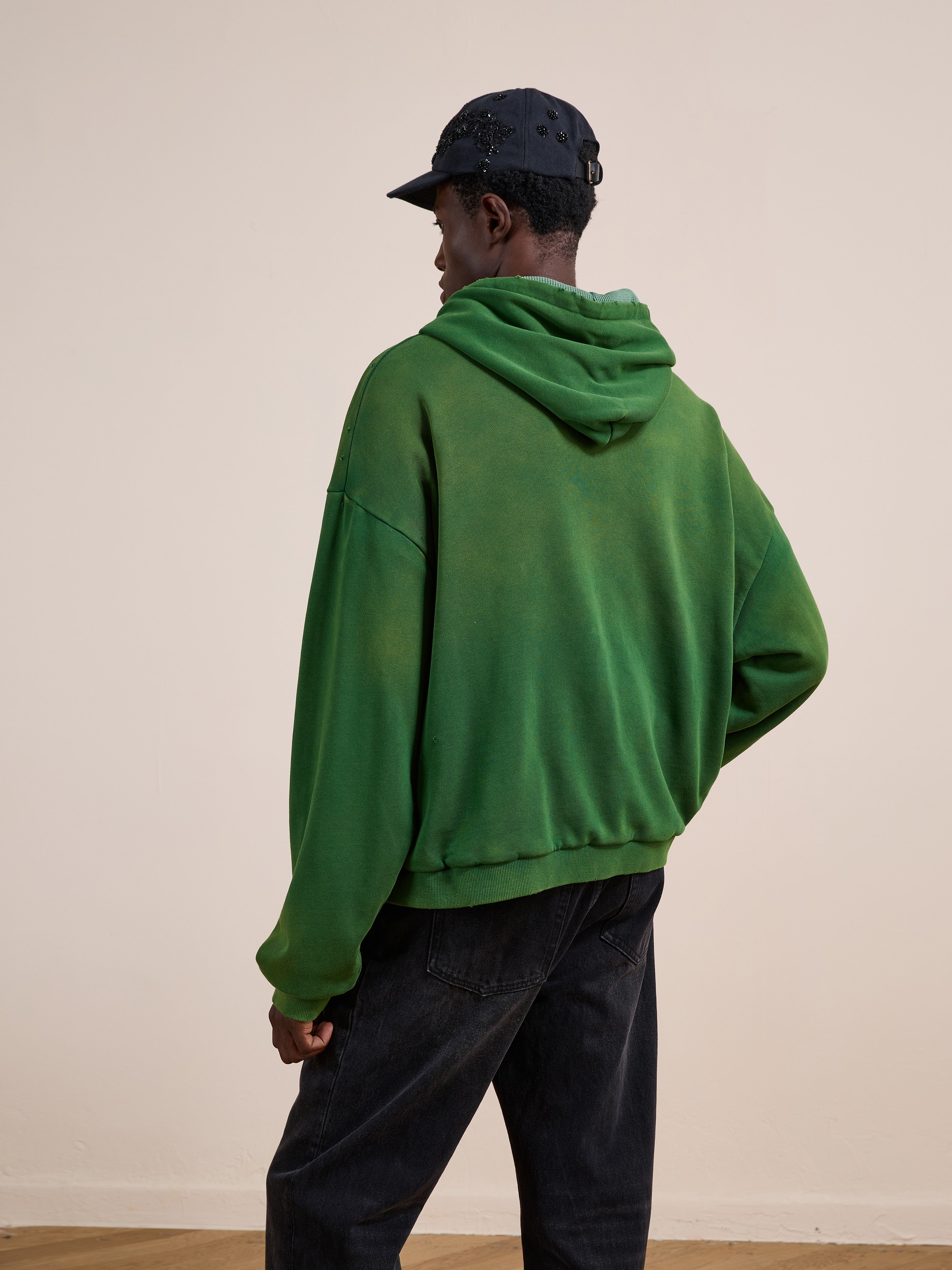 A person wearing a green FOUND Distressed Thermal Lined Zip Up Hoodie, dark jeans, and a dark cap stands indoors facing away from the camera.
