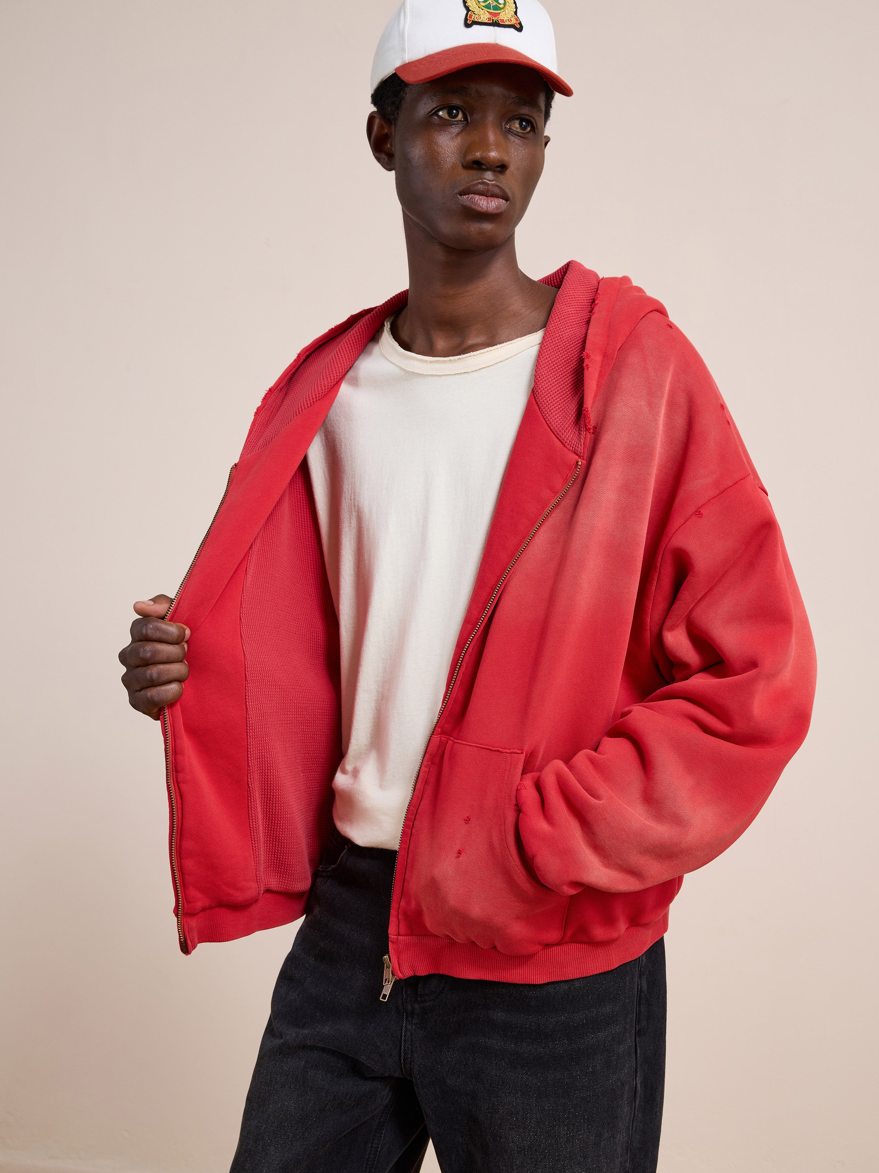 A person in a white t-shirt, black jeans, the FOUND Distressed Thermal Lined Zip Up Hoodie in red (unisex relaxed fit), and a white baseball cap poses against a plain light background.