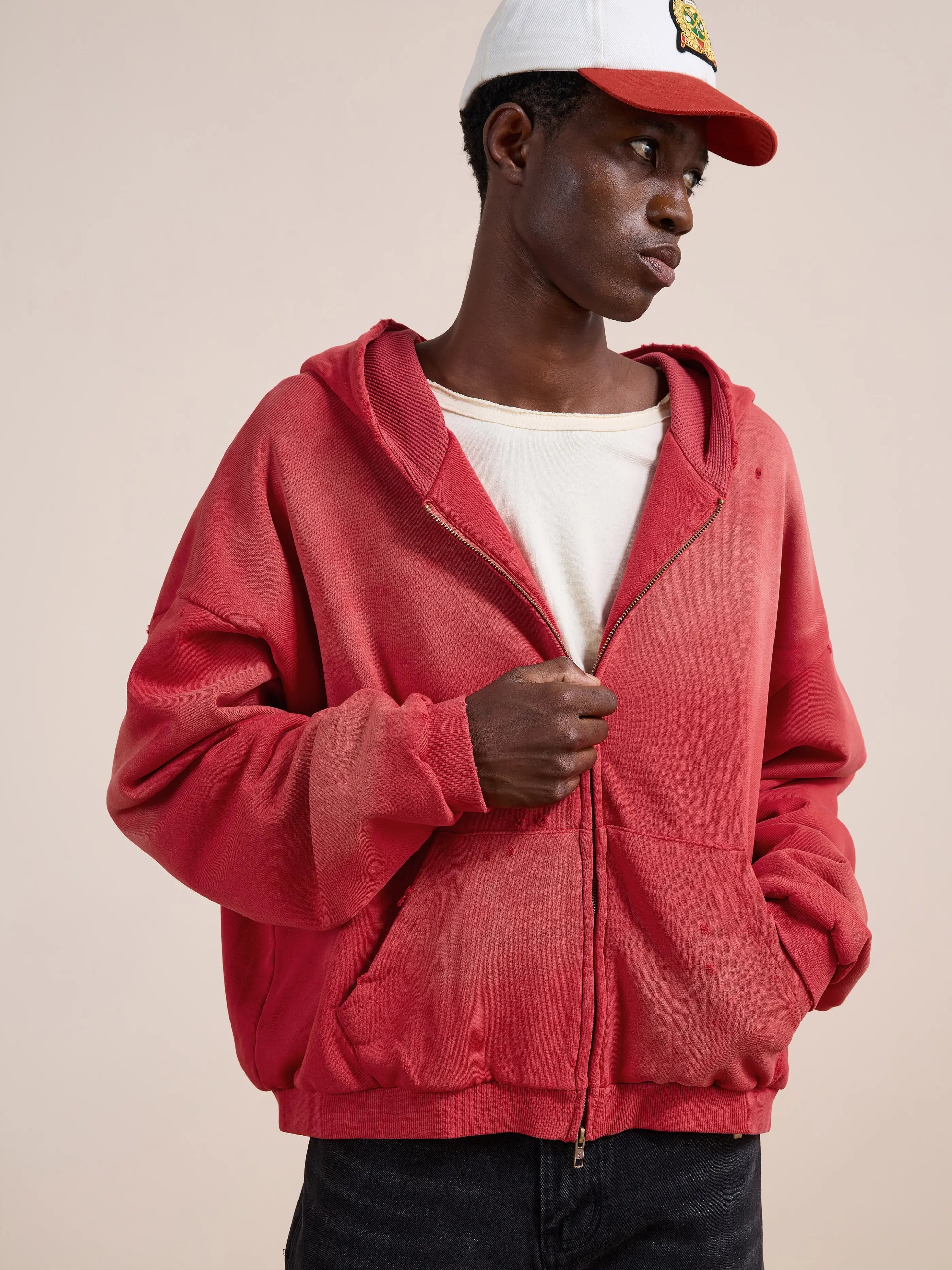 Someone stands against a plain light background wearing a red FOUND Distressed Thermal Lined Zip Up Hoodie, a white t-shirt, and a white baseball cap.