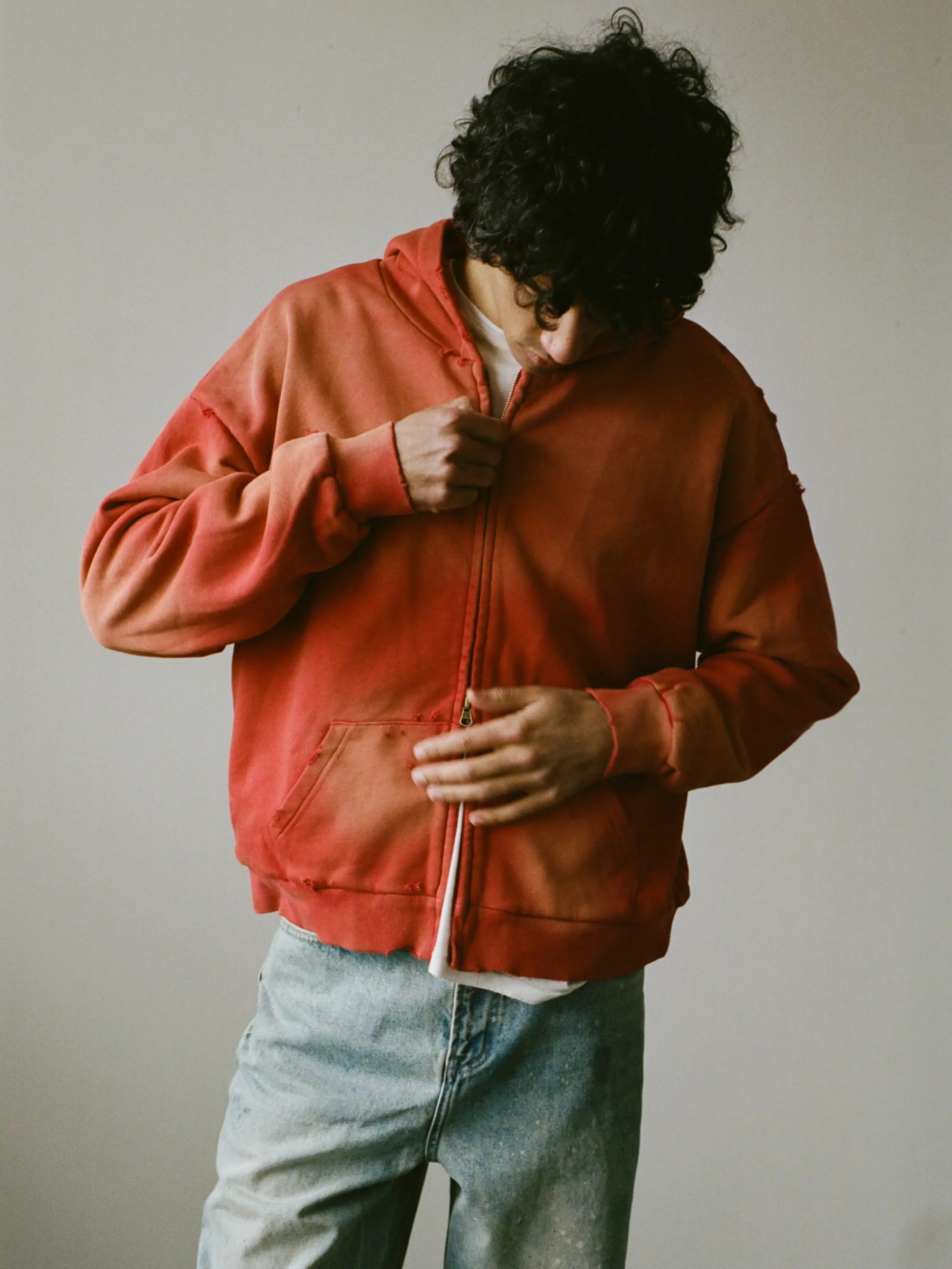 A person with curly hair zips up a red FOUND Distressed Thermal Lined Zip Up Hoodie over a white shirt and light blue jeans, standing against a plain background.