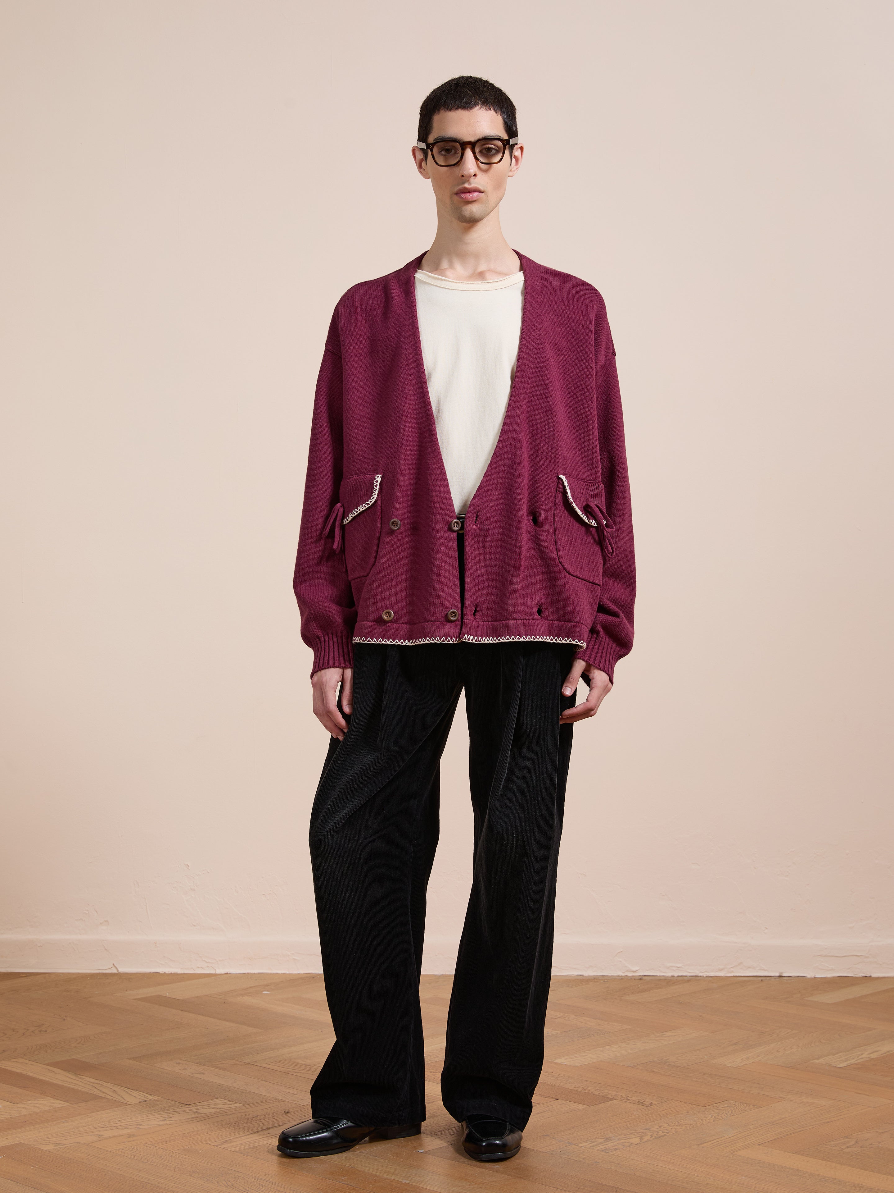 A person stands indoors against a plain light wall wearing glasses, black shoes, black wide-leg pants, a white T-shirt, and the FOUND Double Breasted Bow Cardigan in maroon with pockets.