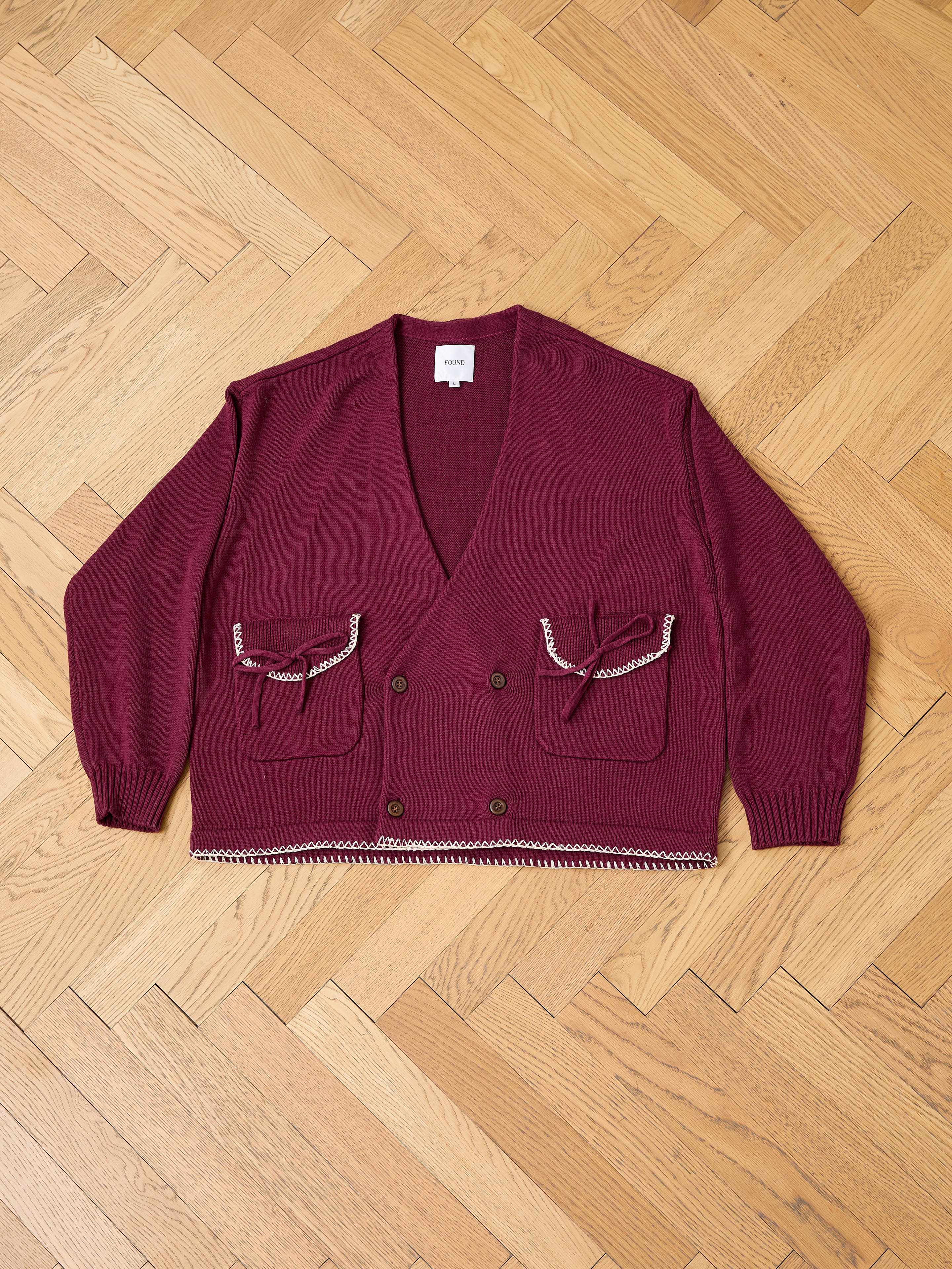 The FOUND Double Breasted Bow Cardigan in maroon features two front pockets with decorative bows and chain trim, displayed flat on a wooden herringbone floor.