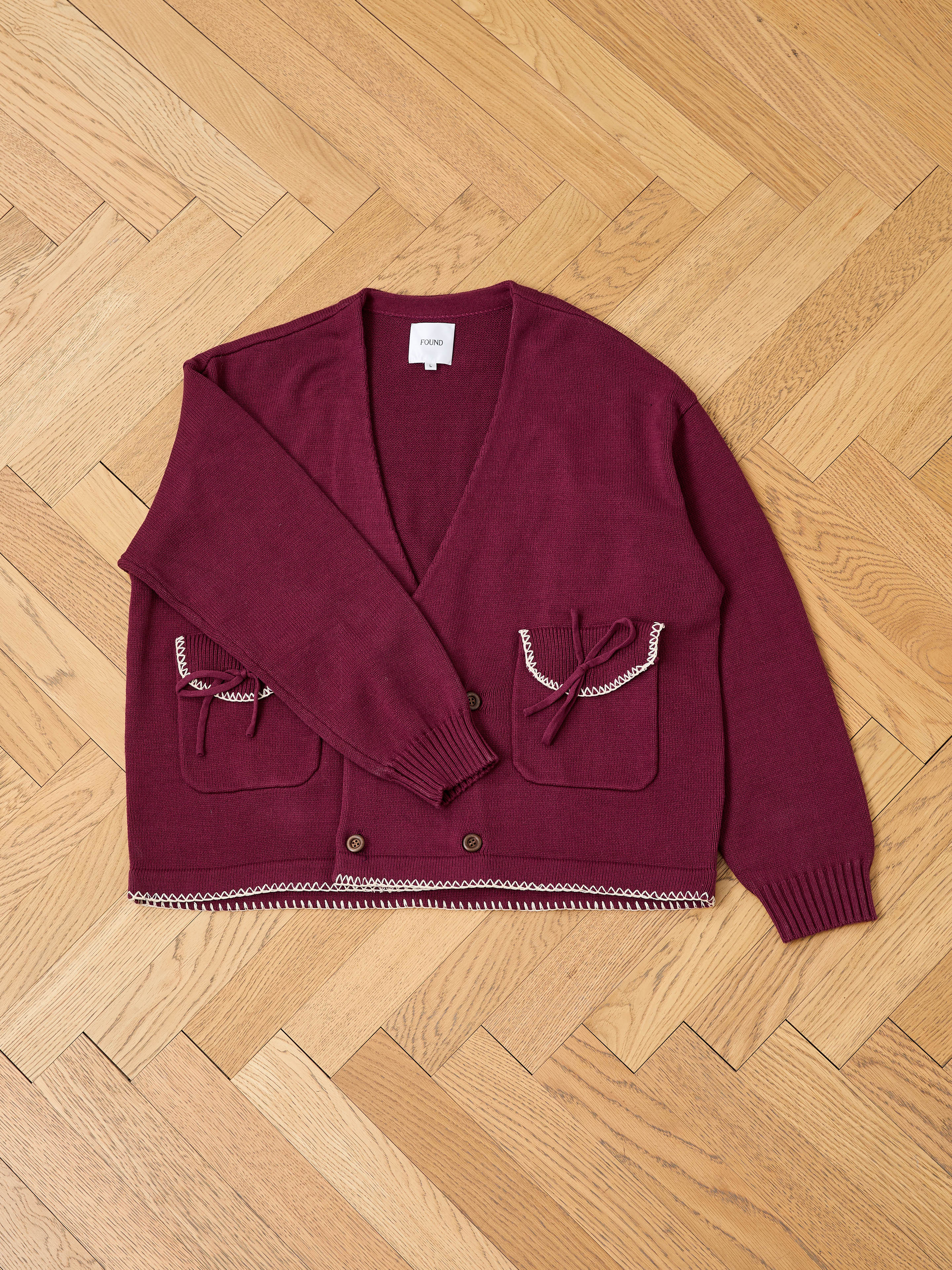 The FOUND Double Breasted Bow Cardigan, a burgundy cotton blend with front pockets, decorative bows, and embroidered trim, is laid flat on a light wood herringbone floor.