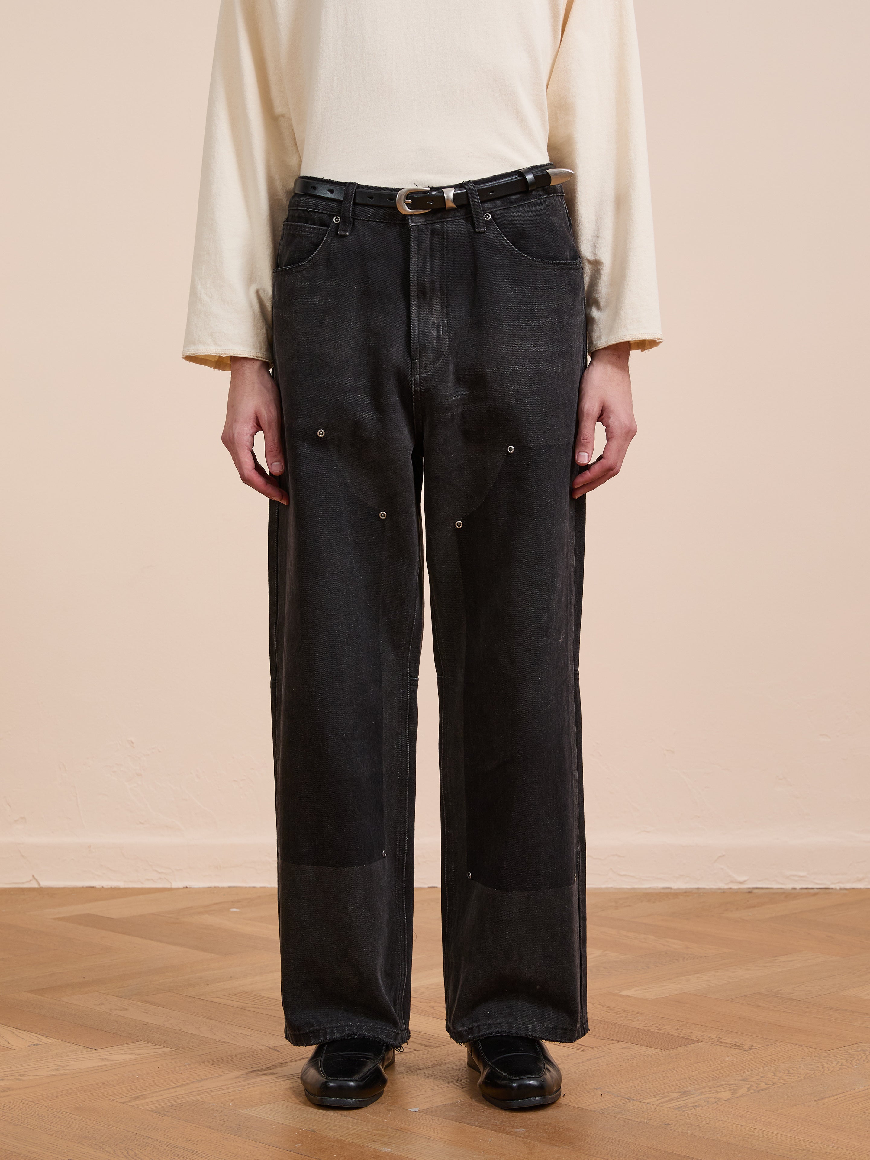 A person wearing a long-sleeve cream shirt and black FOUND Double Knee Carpenter Jeans stands on a wooden floor against a beige wall.