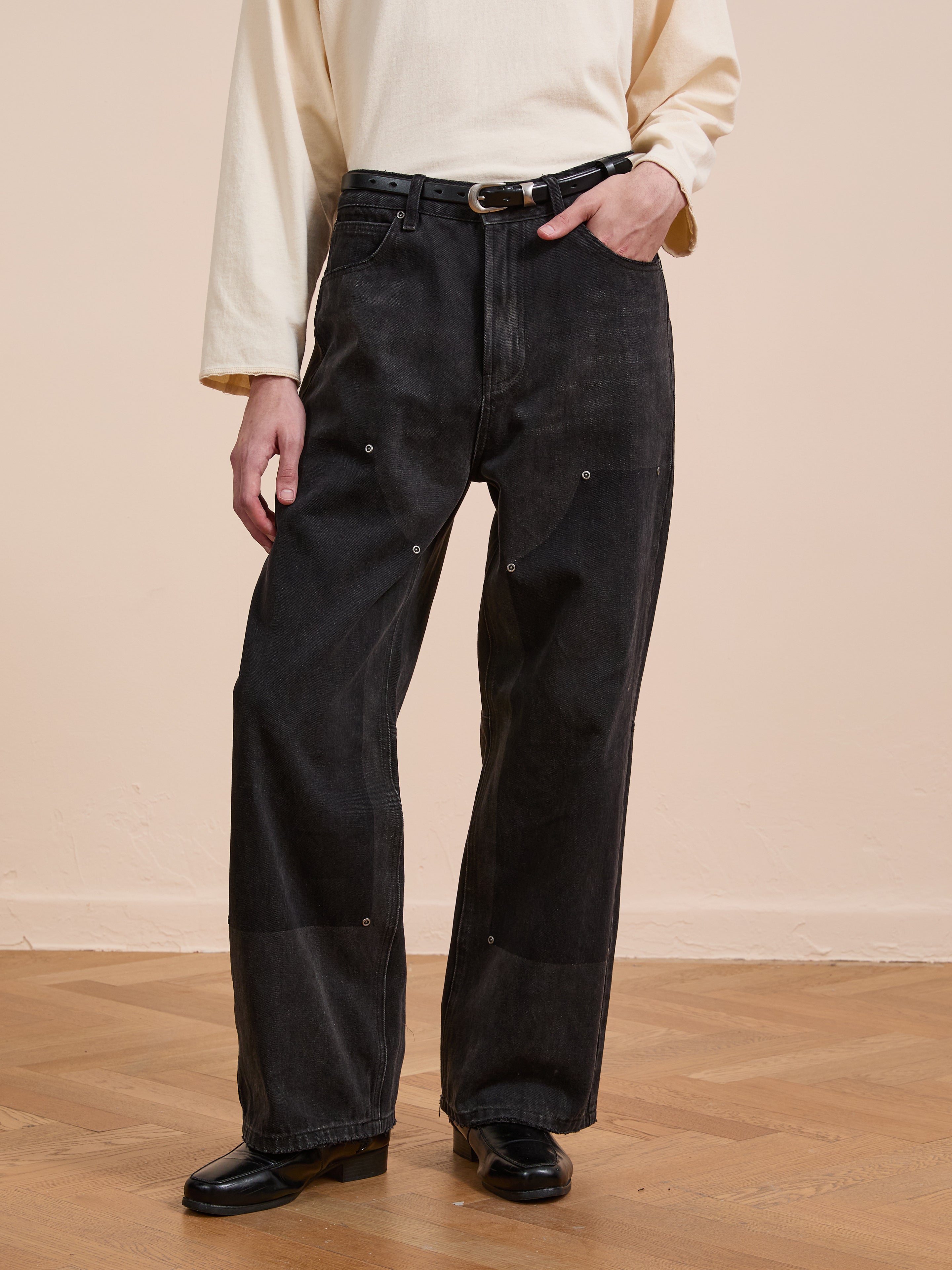 A person wears a long-sleeve cream shirt, FOUND Double Knee Carpenter Jeans in black with decorative stitching and a belt, plus black shoes, standing on a wooden floor against a beige wall.