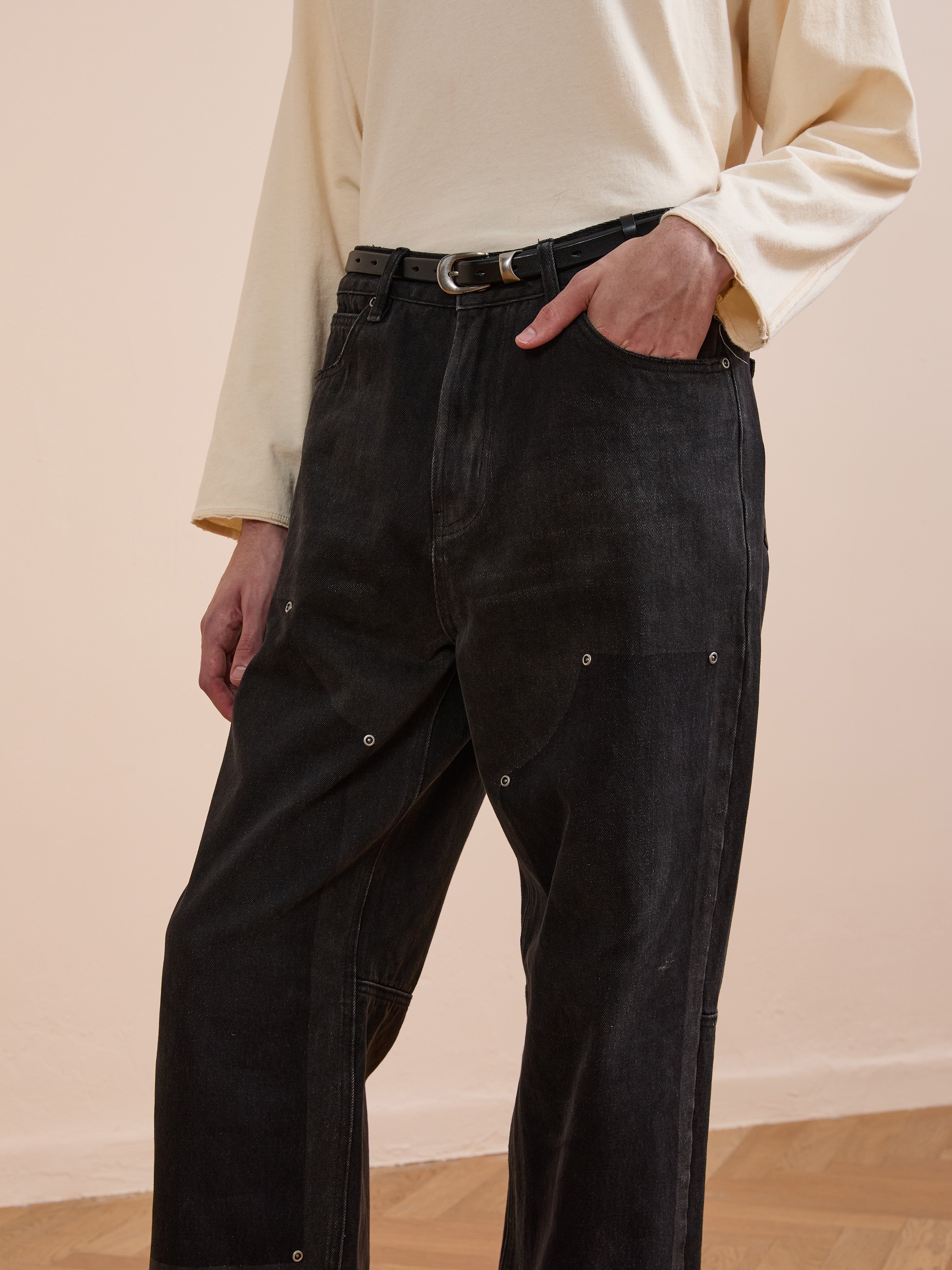 A person wears a long-sleeve beige top tucked into FOUND Double Knee Carpenter Jeans in black, styled with a black belt and standing indoors on a wooden floor.