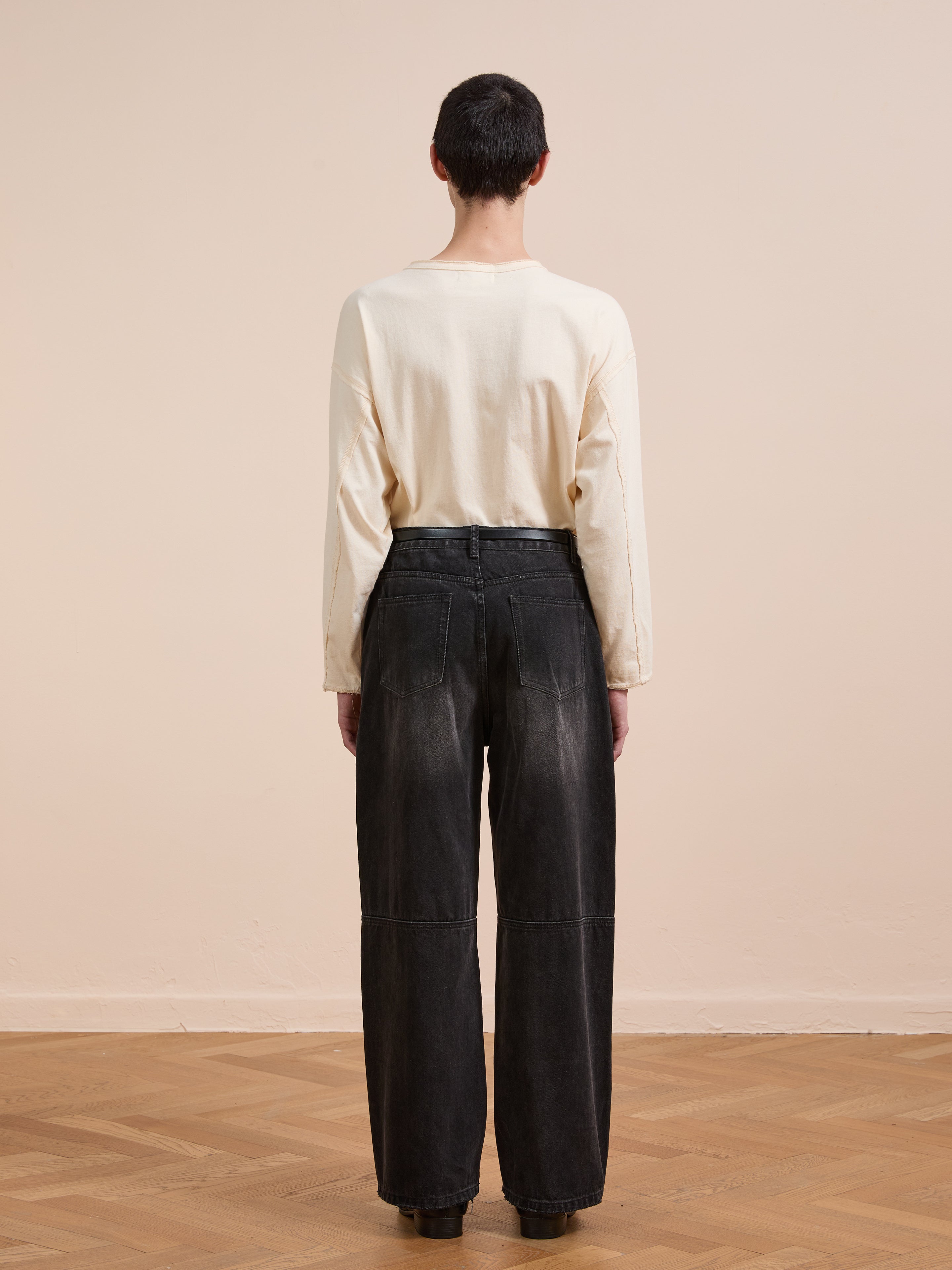 A person with short dark hair stands facing away, wearing a long-sleeve beige shirt and FOUND Double Knee Carpenter Jeans on a wooden floor with a plain beige wall.