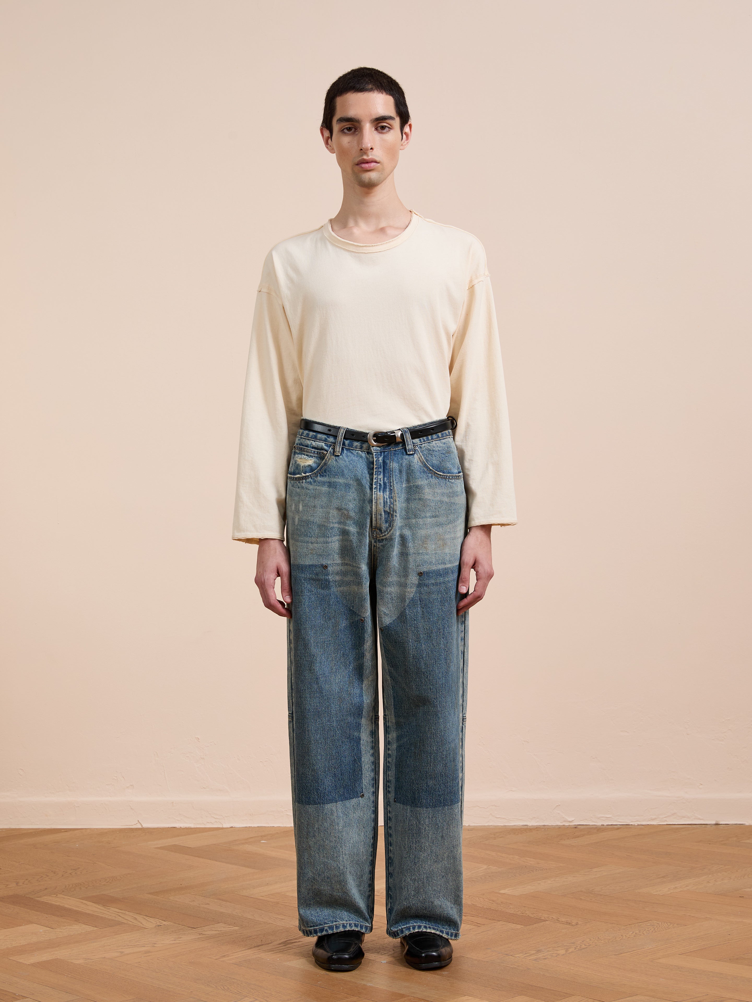 A person stands indoors against a beige wall wearing a loose cream long-sleeve shirt, black shoes, and FOUND Double Knee Carpenter Jeans in a relaxed wide-leg patchwork style.