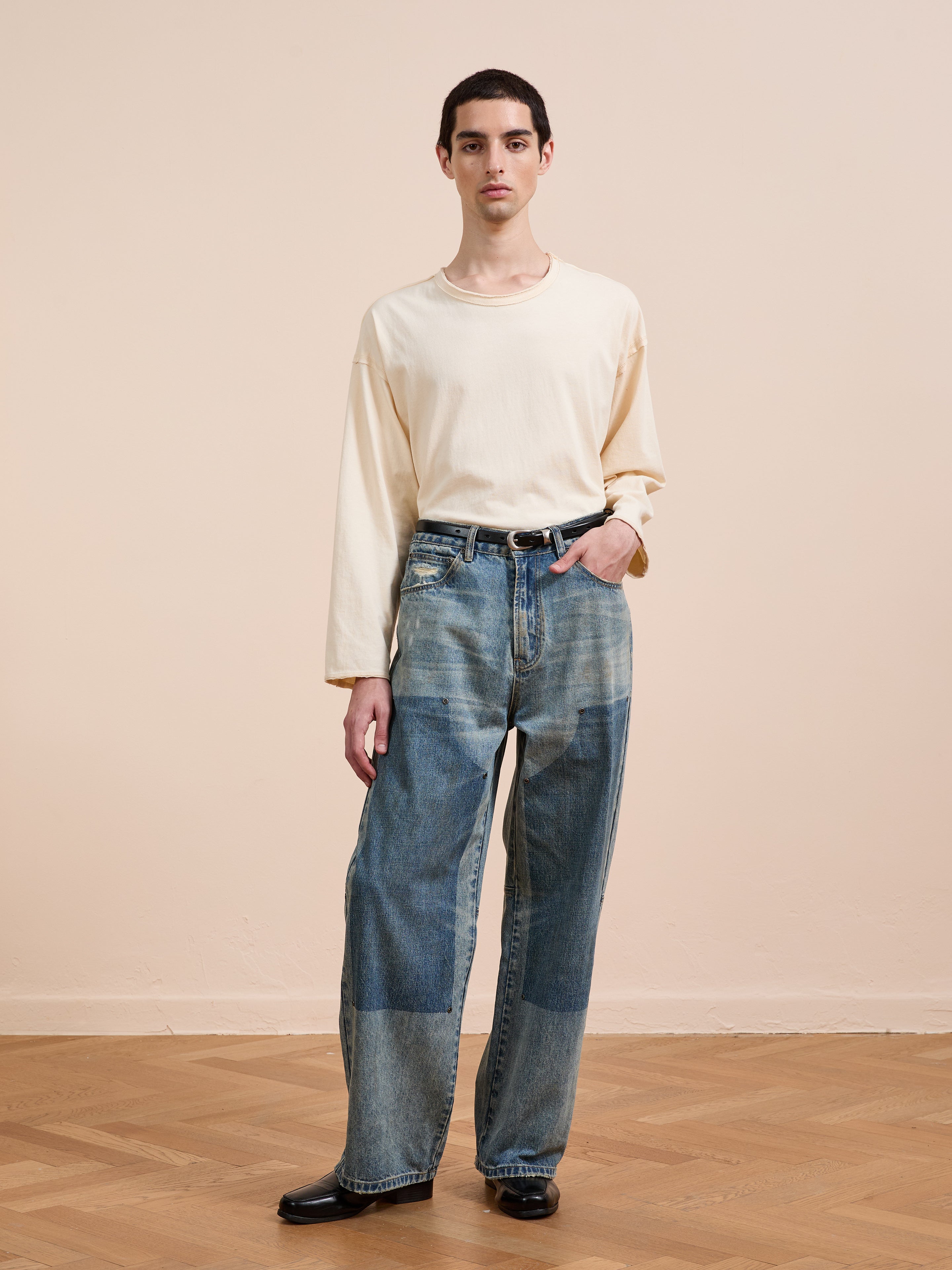 A person stands indoors on a wooden floor against a beige wall, wearing a cream long-sleeve shirt, black shoes, and FOUND Double Knee Carpenter Jeans with patchwork details.