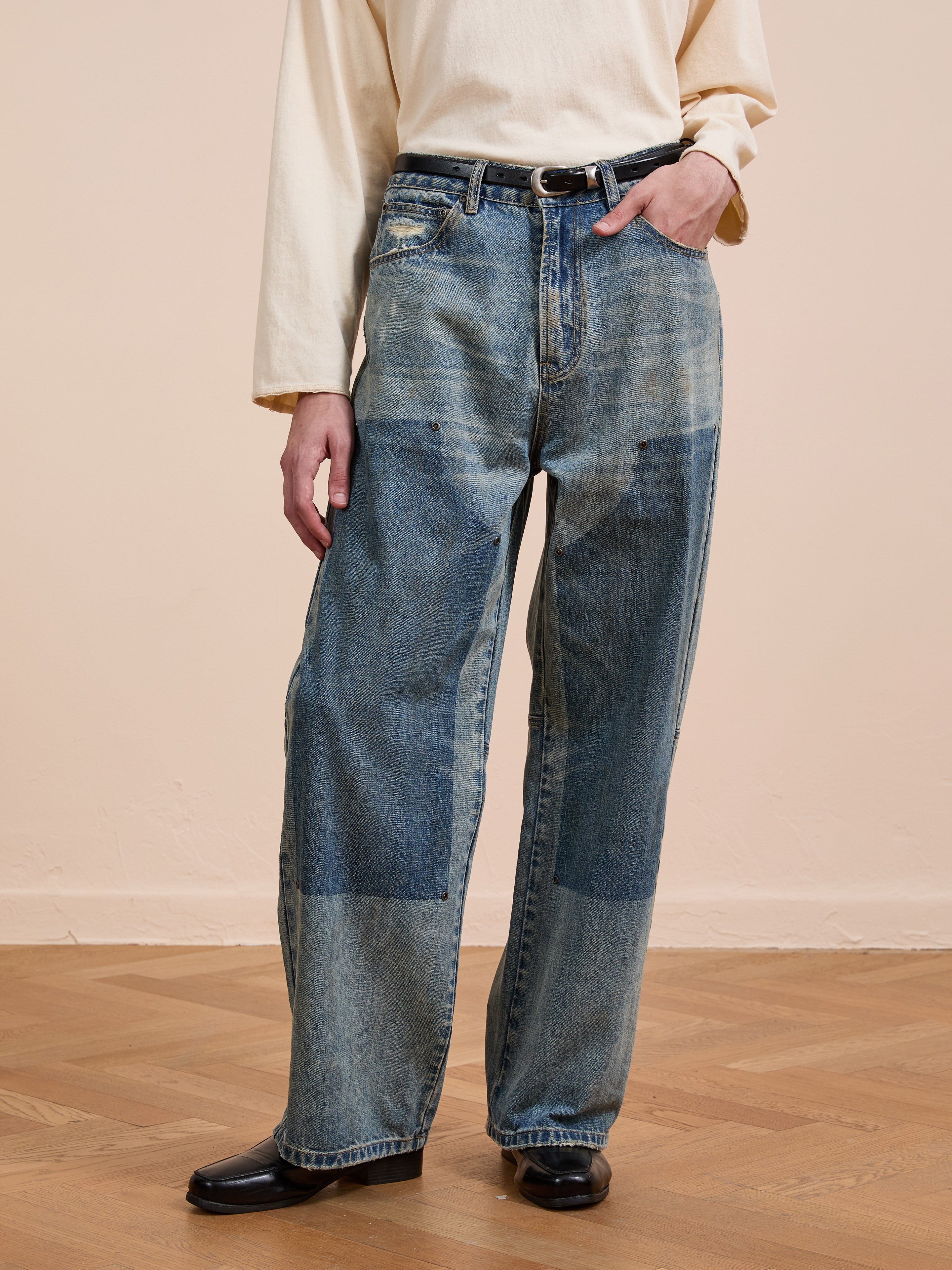 A person wearing FOUND Double Knee Carpenter Jeans in a vintage wash with visible patches, a cream long-sleeve shirt, and black shoes, stands on a wooden floor against a plain wall.