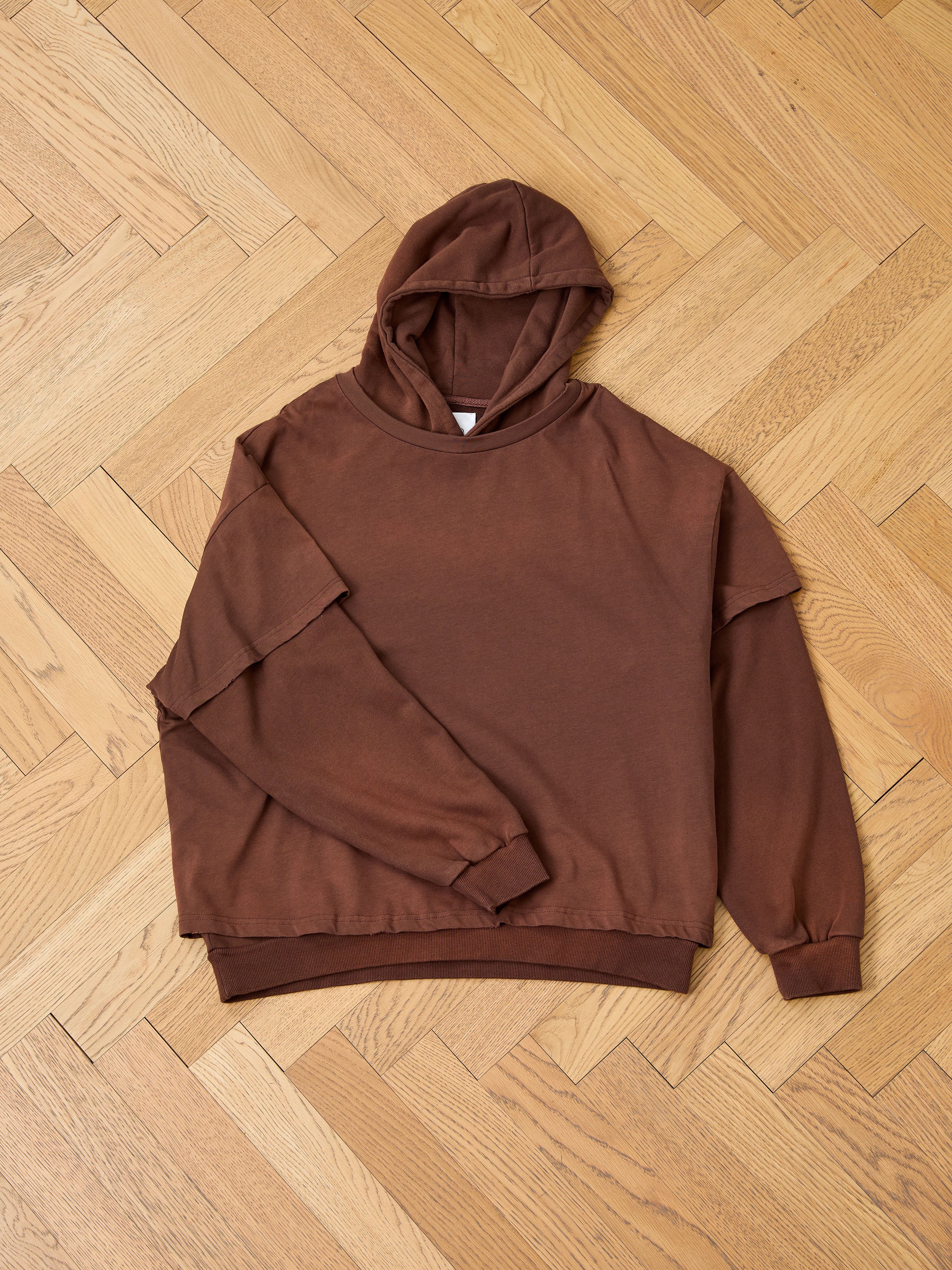 The Found Double Layer Hoodie in brown french terry cotton is displayed flat on a parquet wooden floor, highlighting its cozy vintage style.