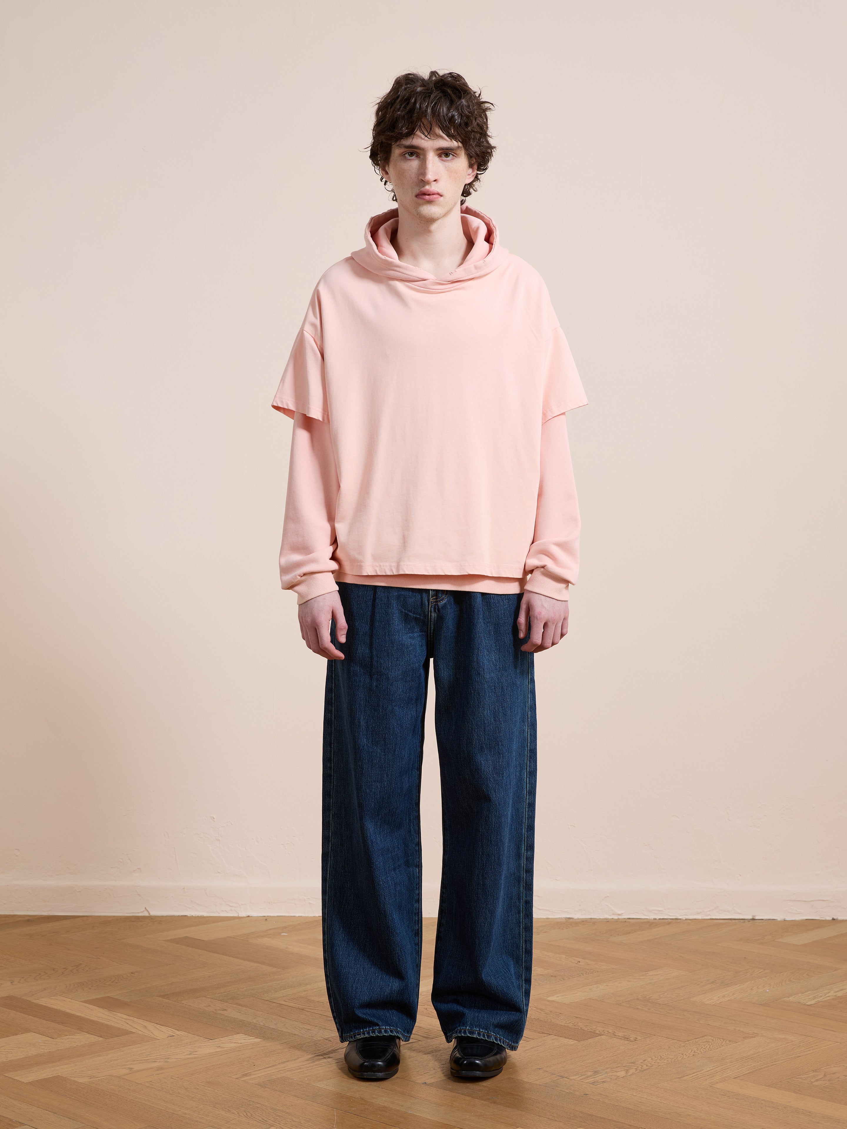 A person stands indoors facing the camera against a beige wall and wooden floor, wearing Found’s Double Layer Hoodie in a relaxed unisex fit (pink French terry cotton) and wide-leg blue jeans.