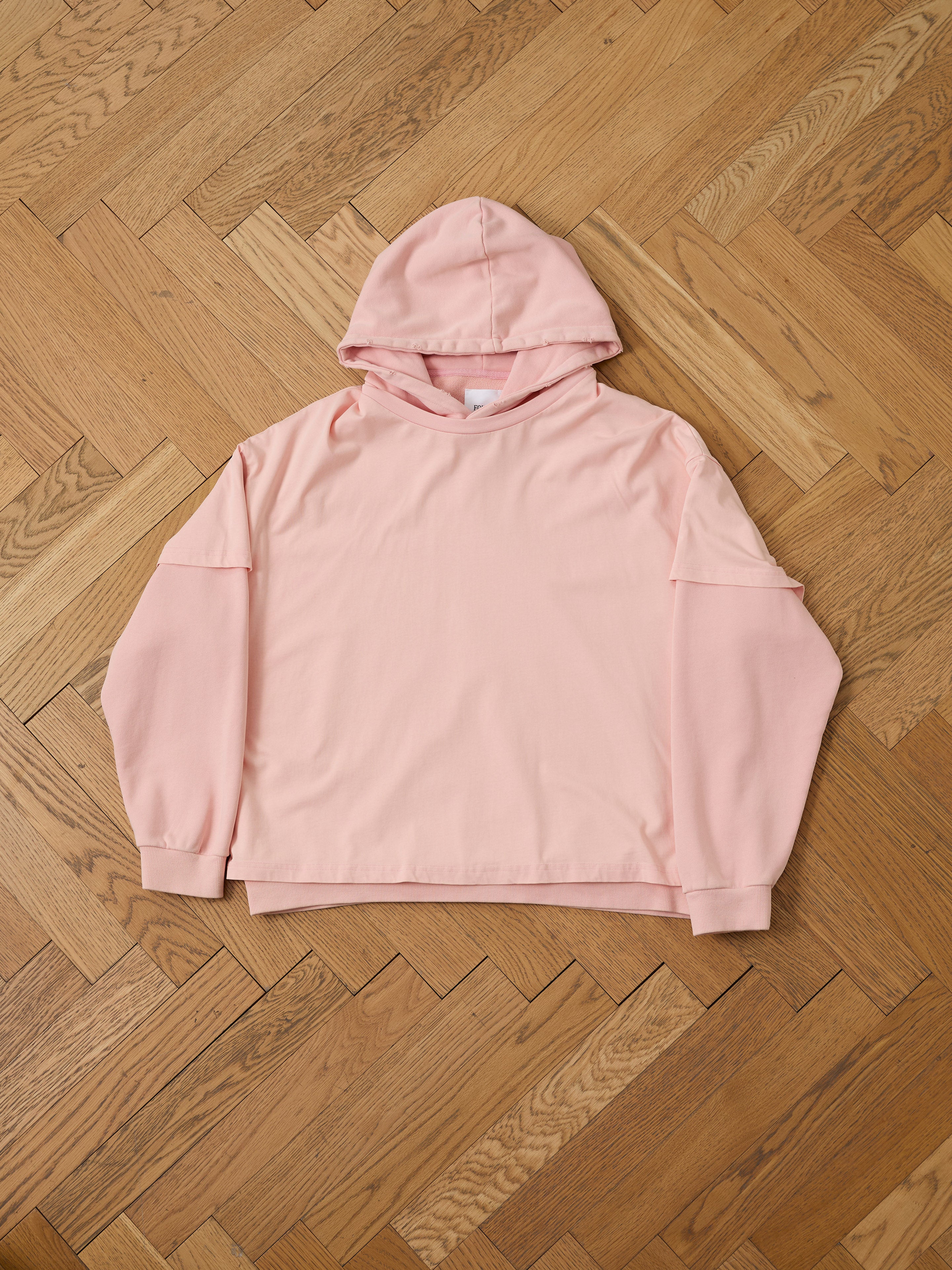 The Found Double Layer Hoodie, a light pink, long-sleeve French terry cotton sweatshirt with a double-layer hood, is laid flat on a wooden parquet floor.