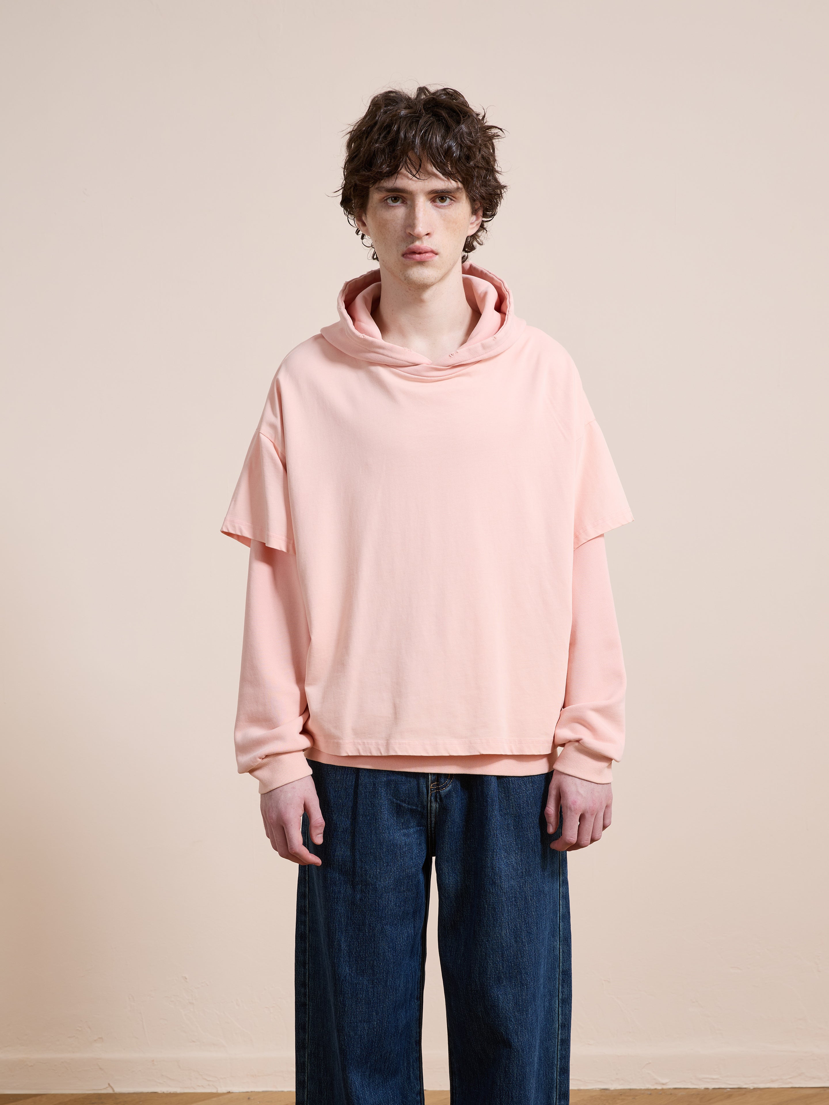 A person with short brown hair wears the Found Double Layer Hoodie in light pink, styled in a loose, unisex relaxed fit, paired with wide-leg blue jeans while standing against a plain beige background.