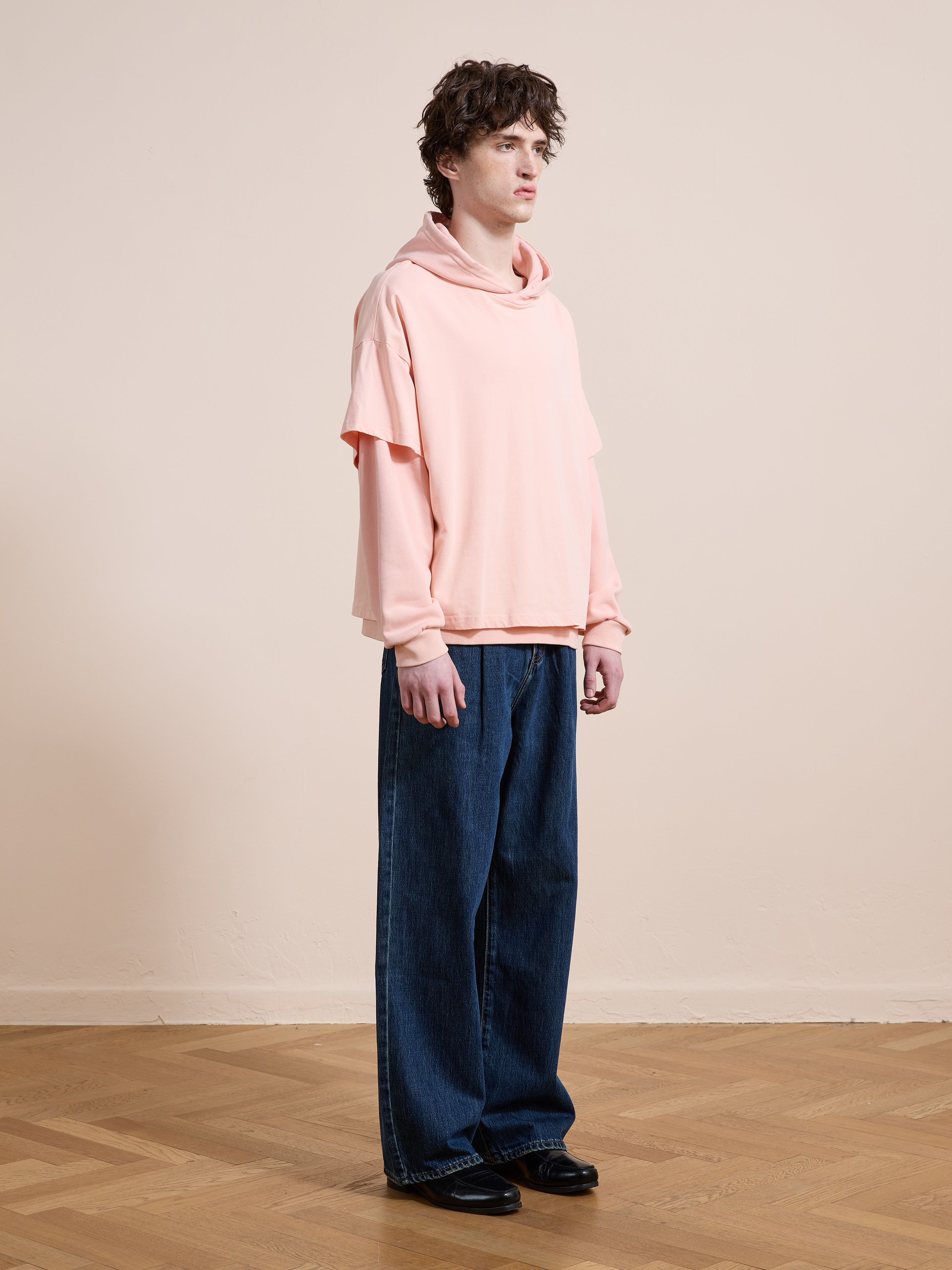 Indoors on a wooden floor, a person faces slightly right wearing Found's Double Layer Hoodie in pink French terry cotton (unisex relaxed fit), wide-leg blue jeans, and black shoes.
