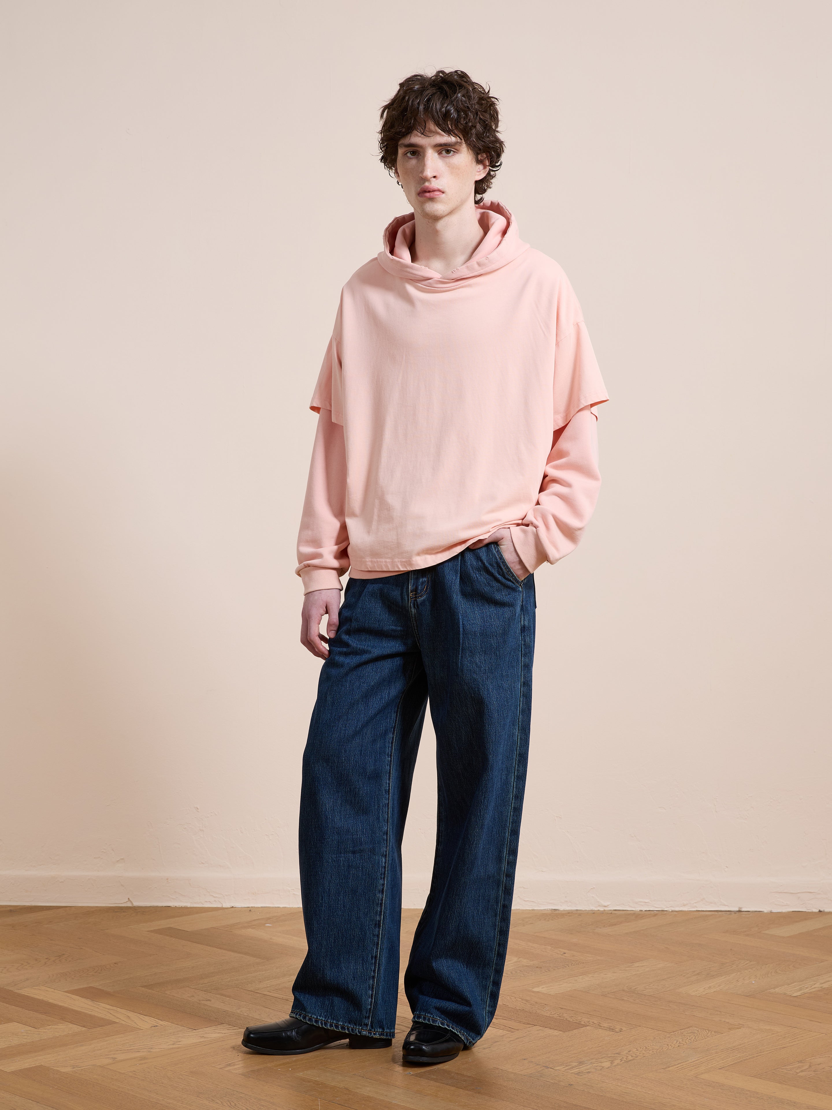 Indoors, a person wears the Found Double Layer Hoodie in pink with a relaxed, unisex fit made from French terry cotton, paired with wide-leg blue jeans and black shoes, one hand slipped into their pocket.