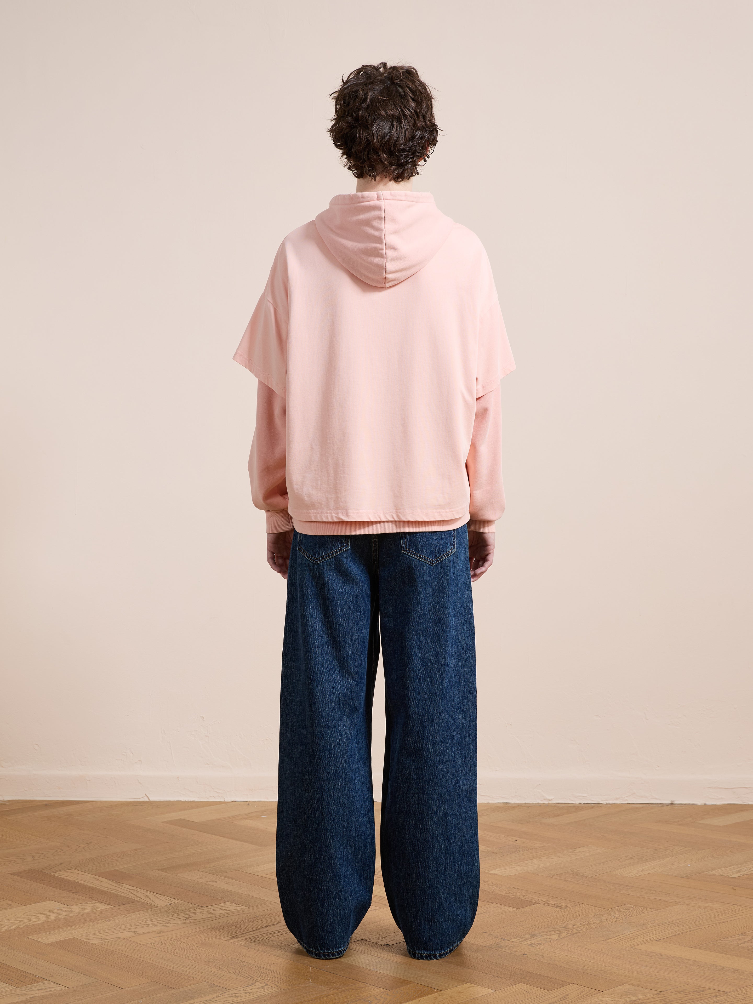 Indoors on a wooden floor, someone faces a plain wall wearing Found’s Double Layer Hoodie in light pink (unisex, relaxed fit) and wide-leg blue jeans.
