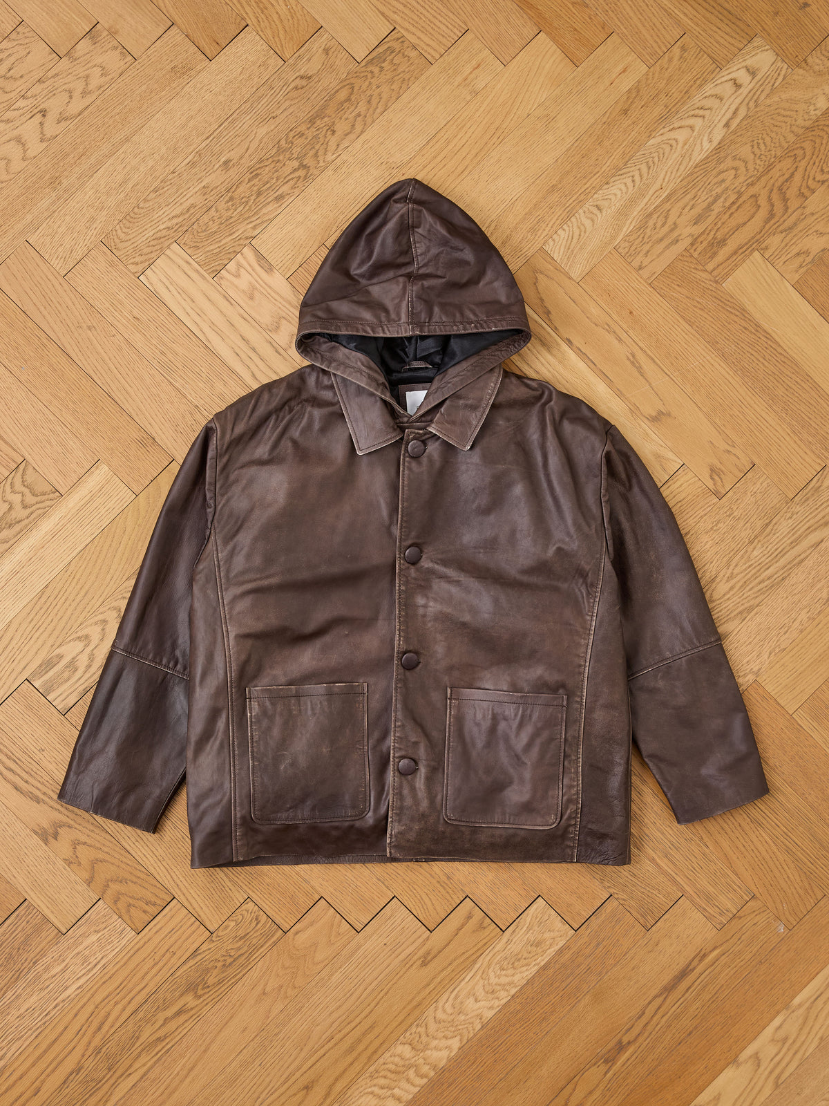 A FOUND Double Layer Leather Hooded Jacket in brown, designed for unisex wear, featuring front buttons and two large front pockets, is displayed laid flat on a wooden herringbone-patterned floor.