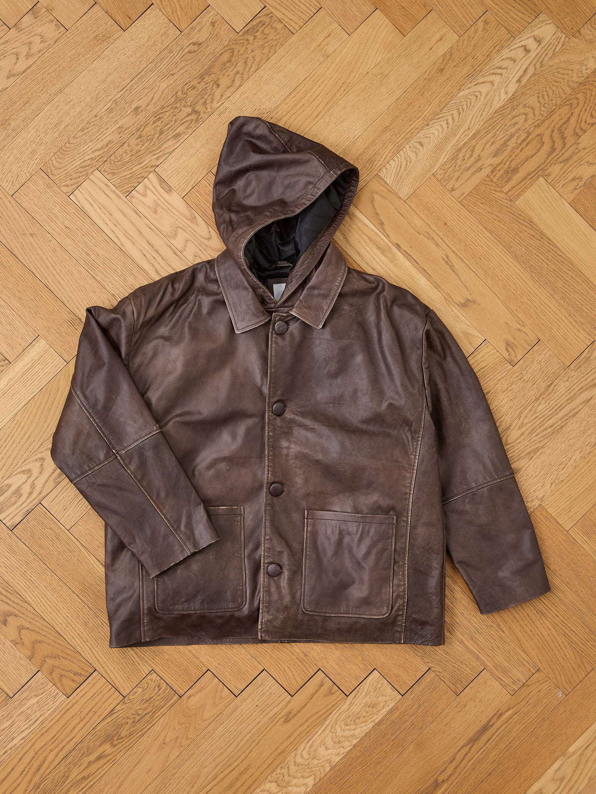 FOUND Double Layer Leather Hooded Jacket in brown leather, featuring a hood, front button closure, and spacious pockets, laid flat on a wooden herringbone-pattern floor. This unisex jacket offers a comfortable fit and timeless style for versatile everyday wear.