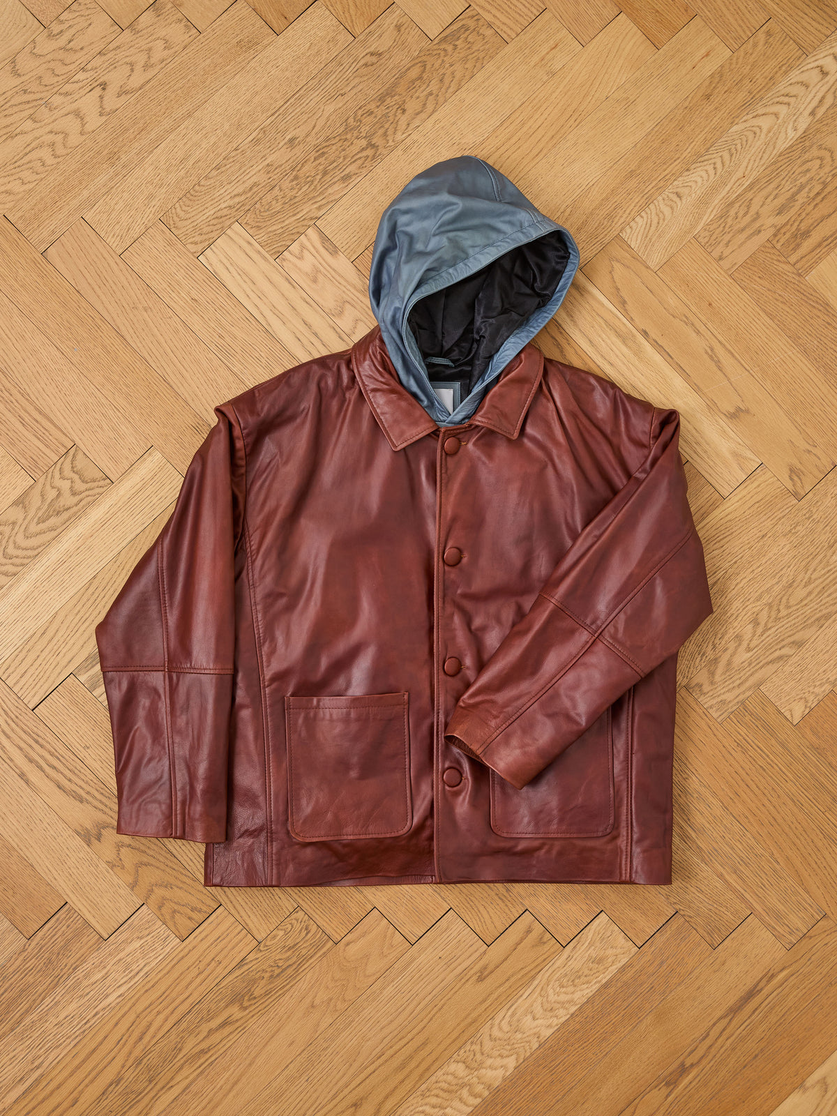 The FOUND Double Layer Leather Hooded Jacket, a brown unisex leather jacket with large buttons, front pockets, and a contrasting blue-gray hood, is displayed flat on a light wood herringbone floor.