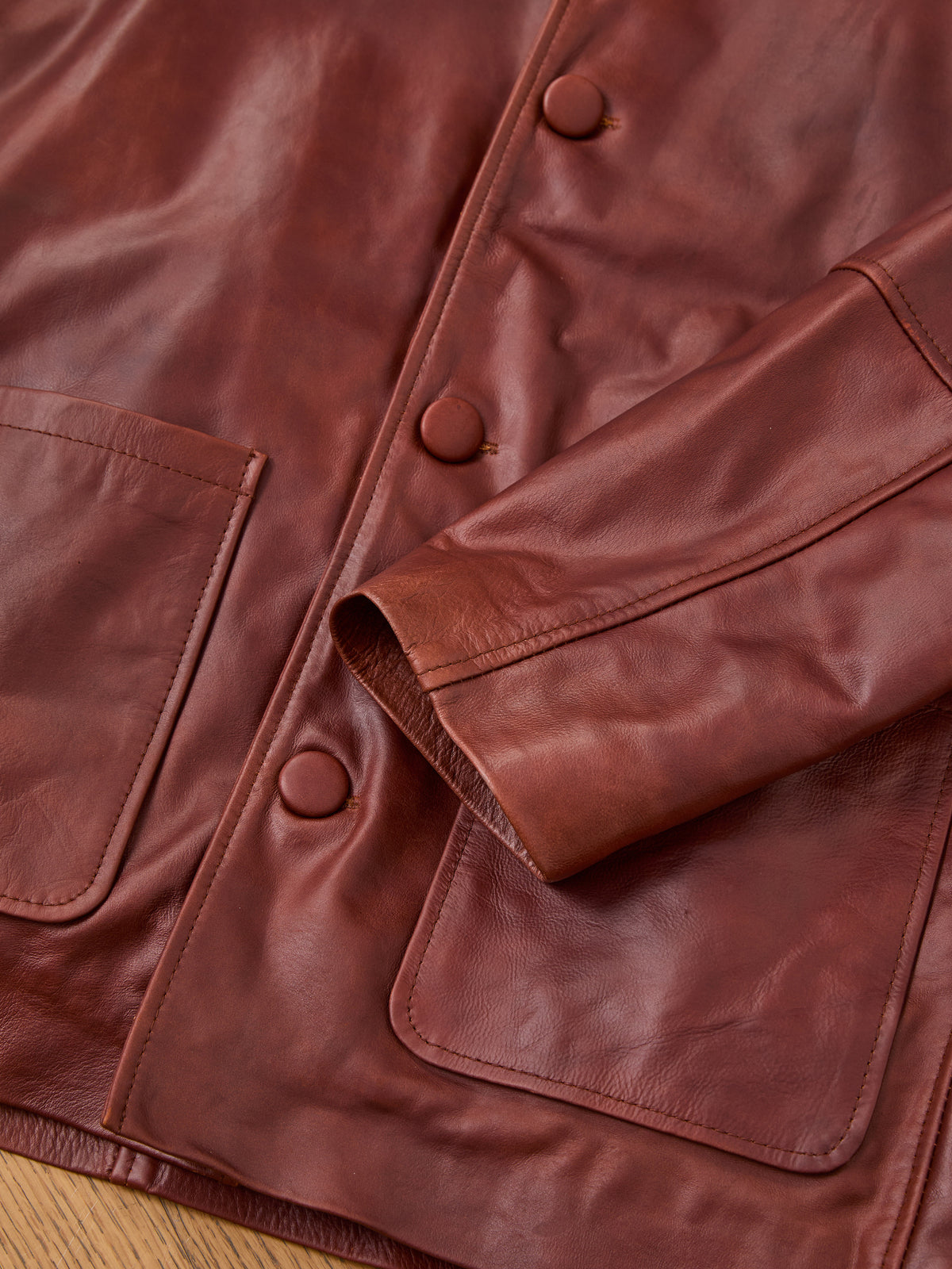 Close-up of the FOUND Double Layer Leather Hooded Jacket—an effortlessly stylish unisex outerwear piece, featuring large pockets and covered buttons, displayed on a wooden surface.