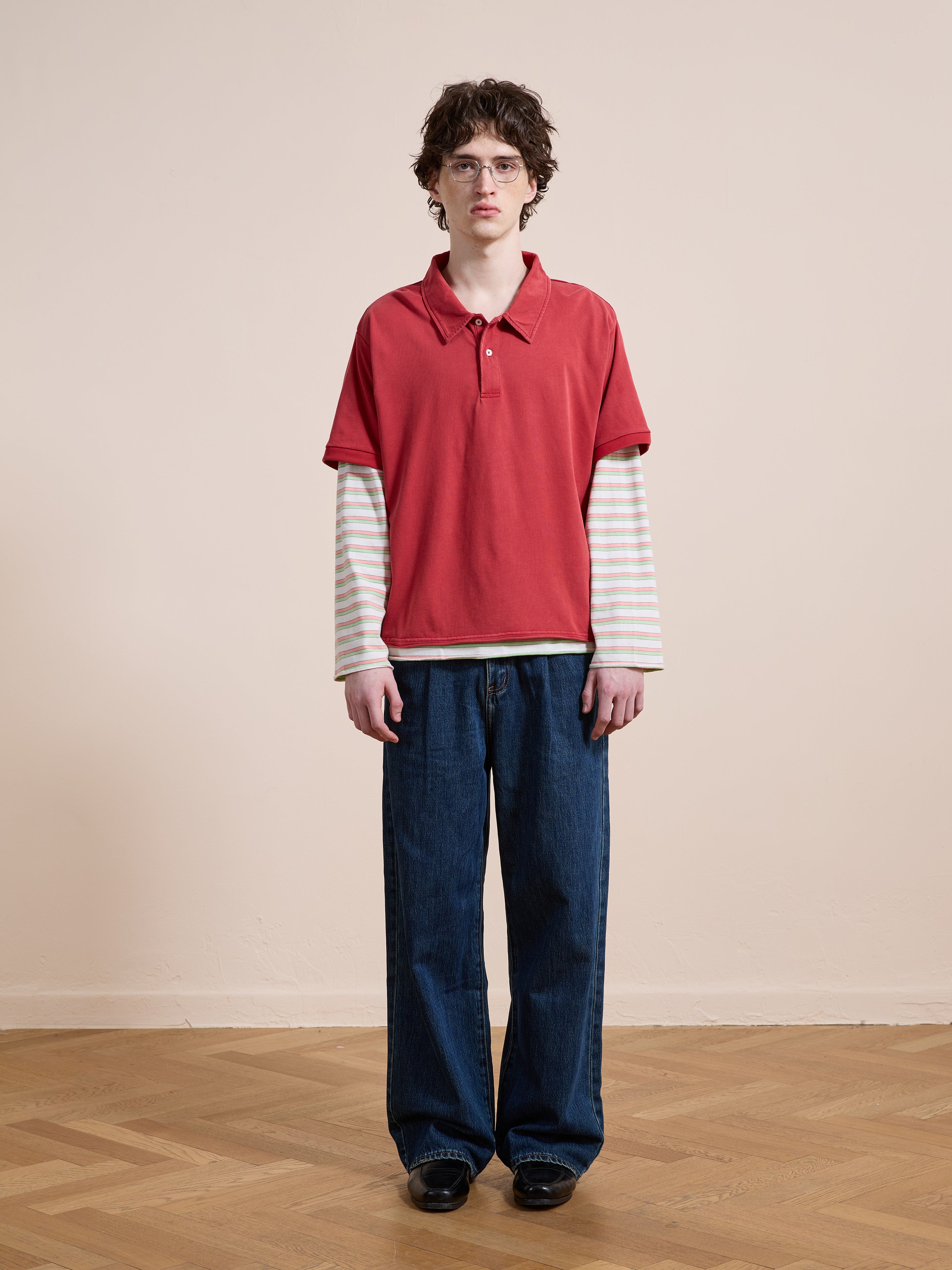 Indoors on a wooden floor, someone wears FOUND’s Double Layer Stripe Polo in a unisex relaxed fit with wide blue jeans and black shoes against a plain beige wall, channeling retro athletic sportswear style.
