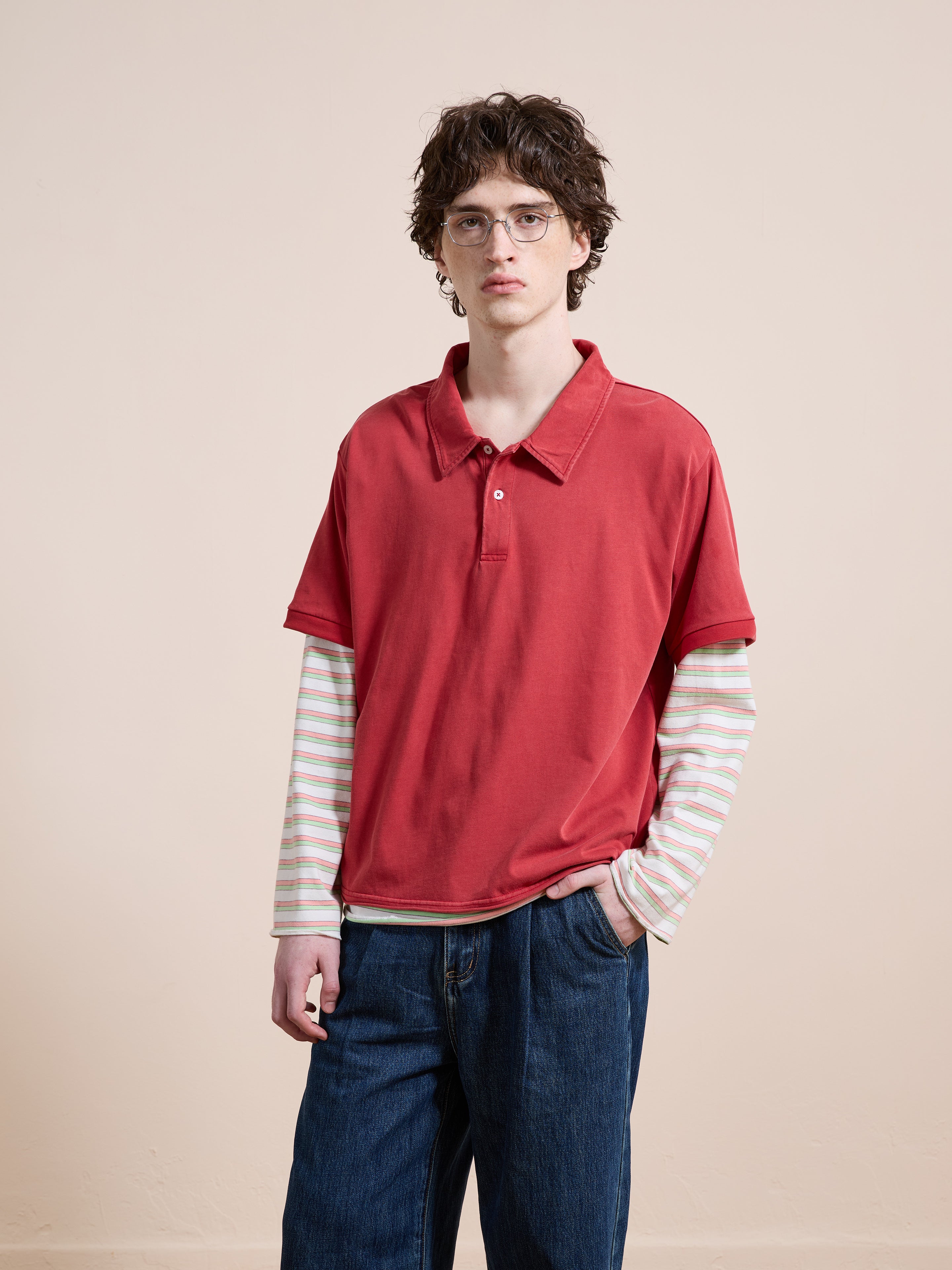 A person models the FOUND Double Layer Stripe Polo—red over striped long sleeves—for a unisex, relaxed-fit vintage sportswear look with blue jeans against a plain beige background.