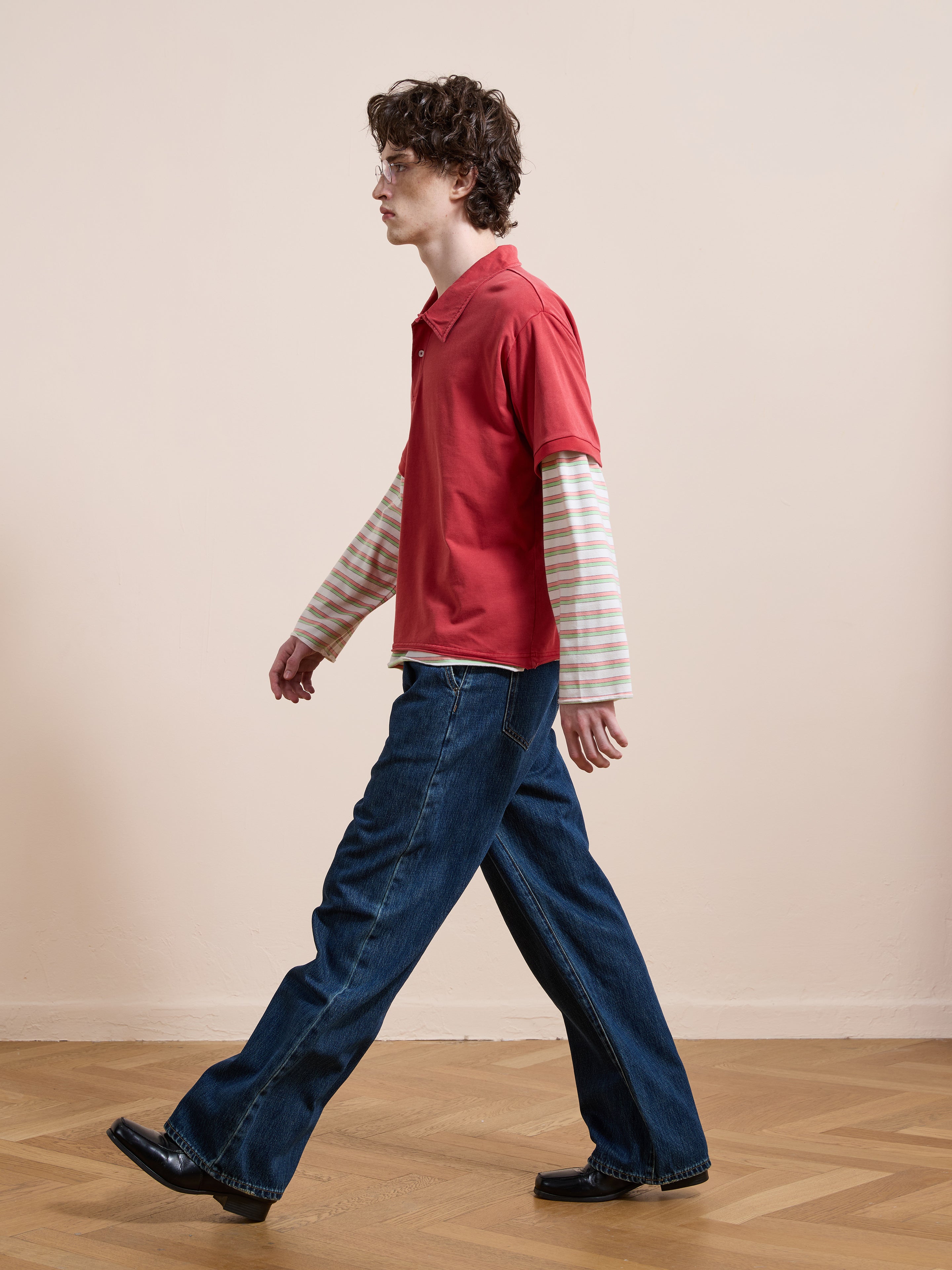 A person with curly hair and glasses walks indoors on a wooden floor wearing blue jeans, black shoes, and the FOUND Double Layer Stripe Polo—a vintage athletic sportswear-inspired red shirt layered over stripes.