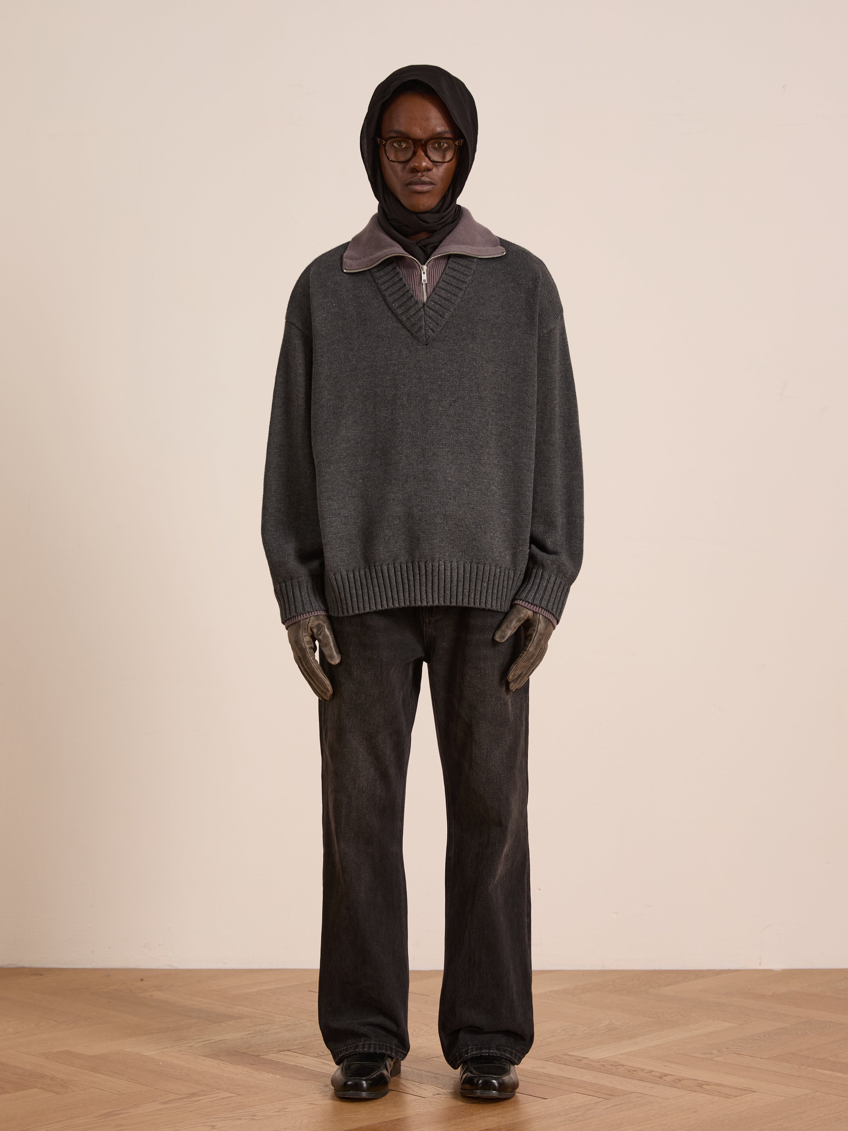 A person stands against a plain wall wearing FOUND’s Double Layer Sweater—a wool blend gray pullover over a dark hoodie, paired with wide-leg black pants, gloves, glasses, and black shoes—ideal for cold-weather layering.