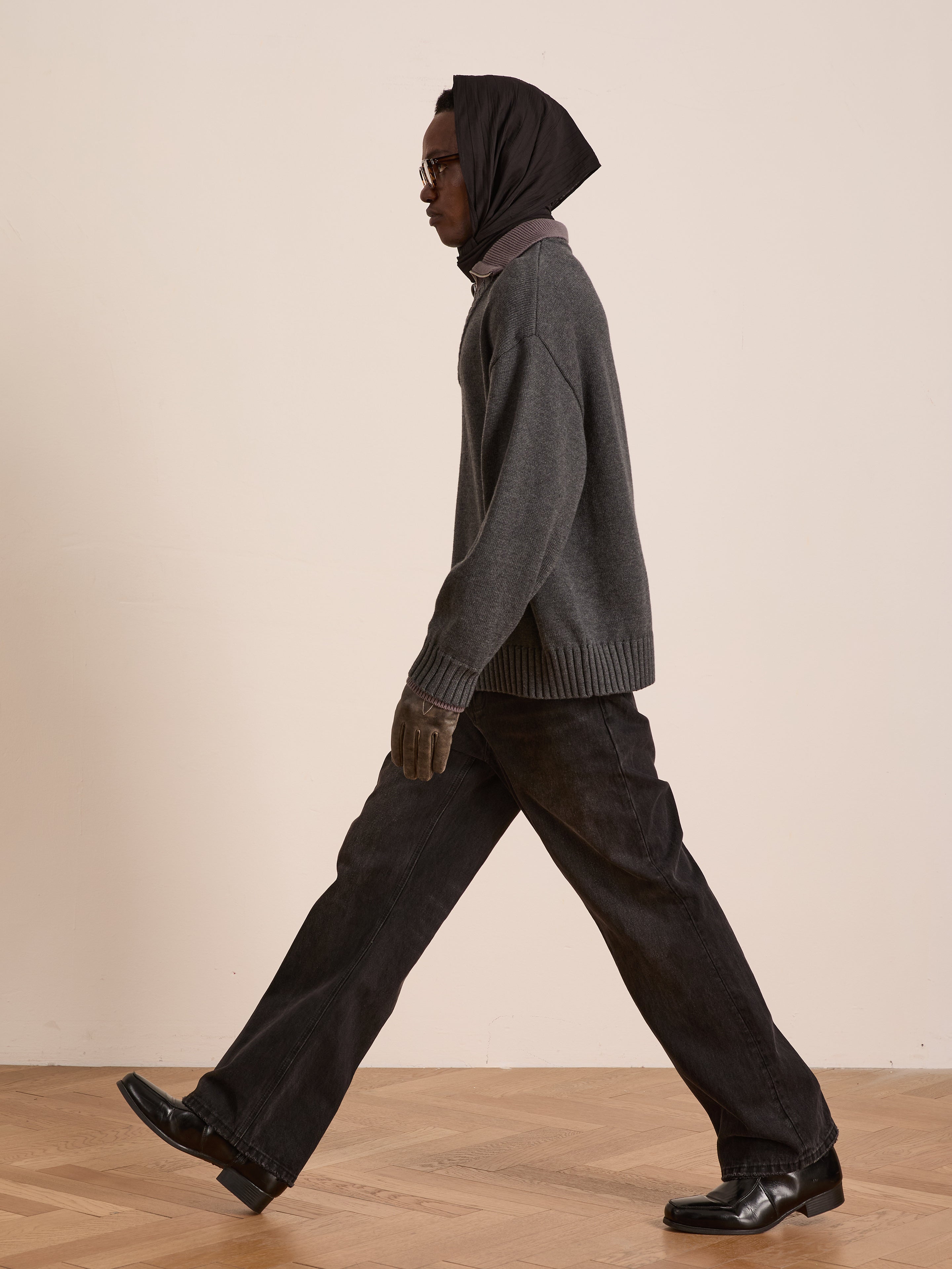 Wearing the FOUND Double Layer Sweater, black headscarf, glasses, gloves, dark pants, and black shoes, a person walks indoors on a wooden floor—showcasing stylish cold-weather layering.