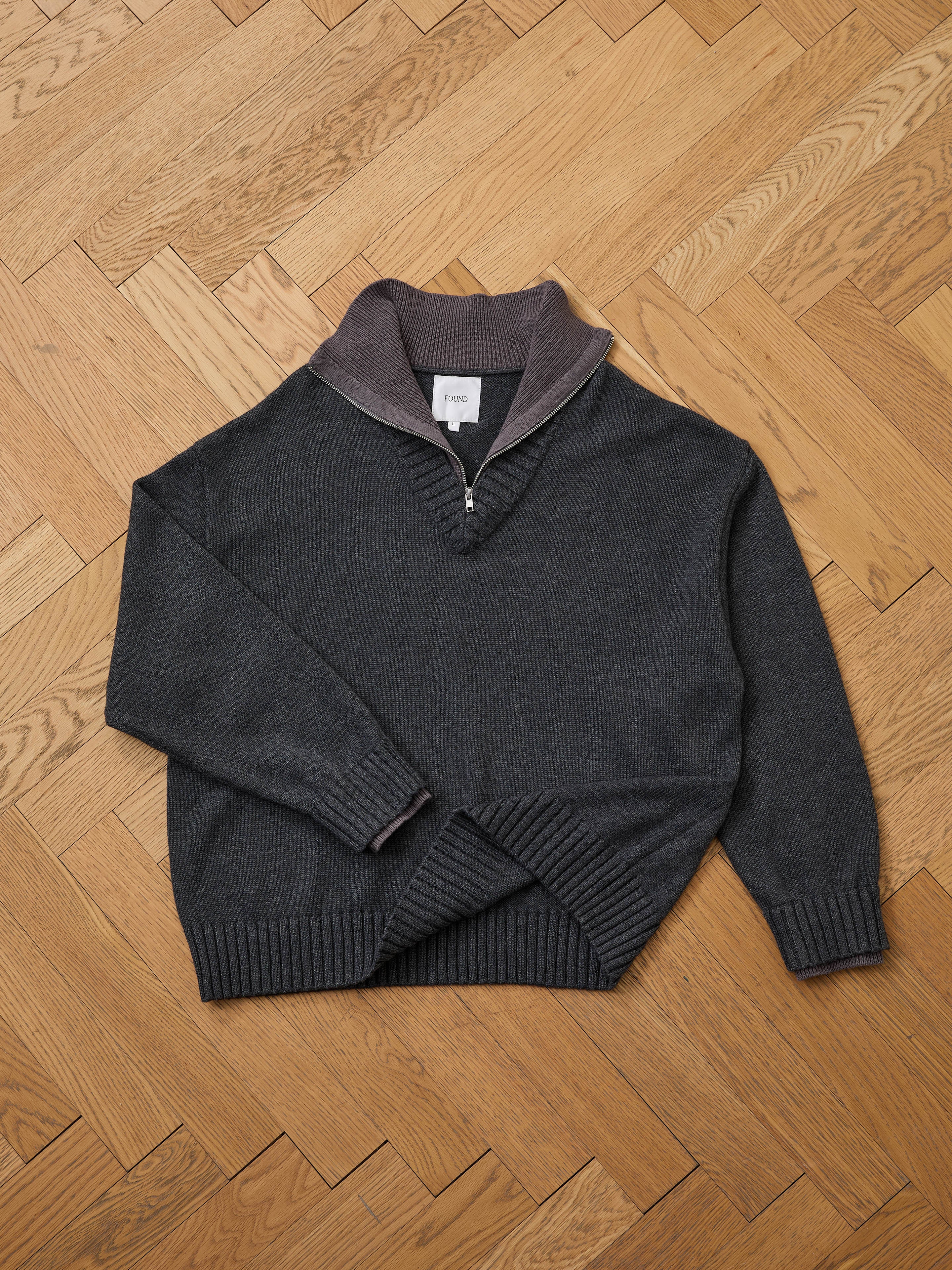 The FOUND Double Layer Sweater, featuring a partial zipper and ribbed cuffs, hem, and collar in dark gray wool blend, is shown lying flat on a wooden parquet floor—ideal for layering in cold weather.
