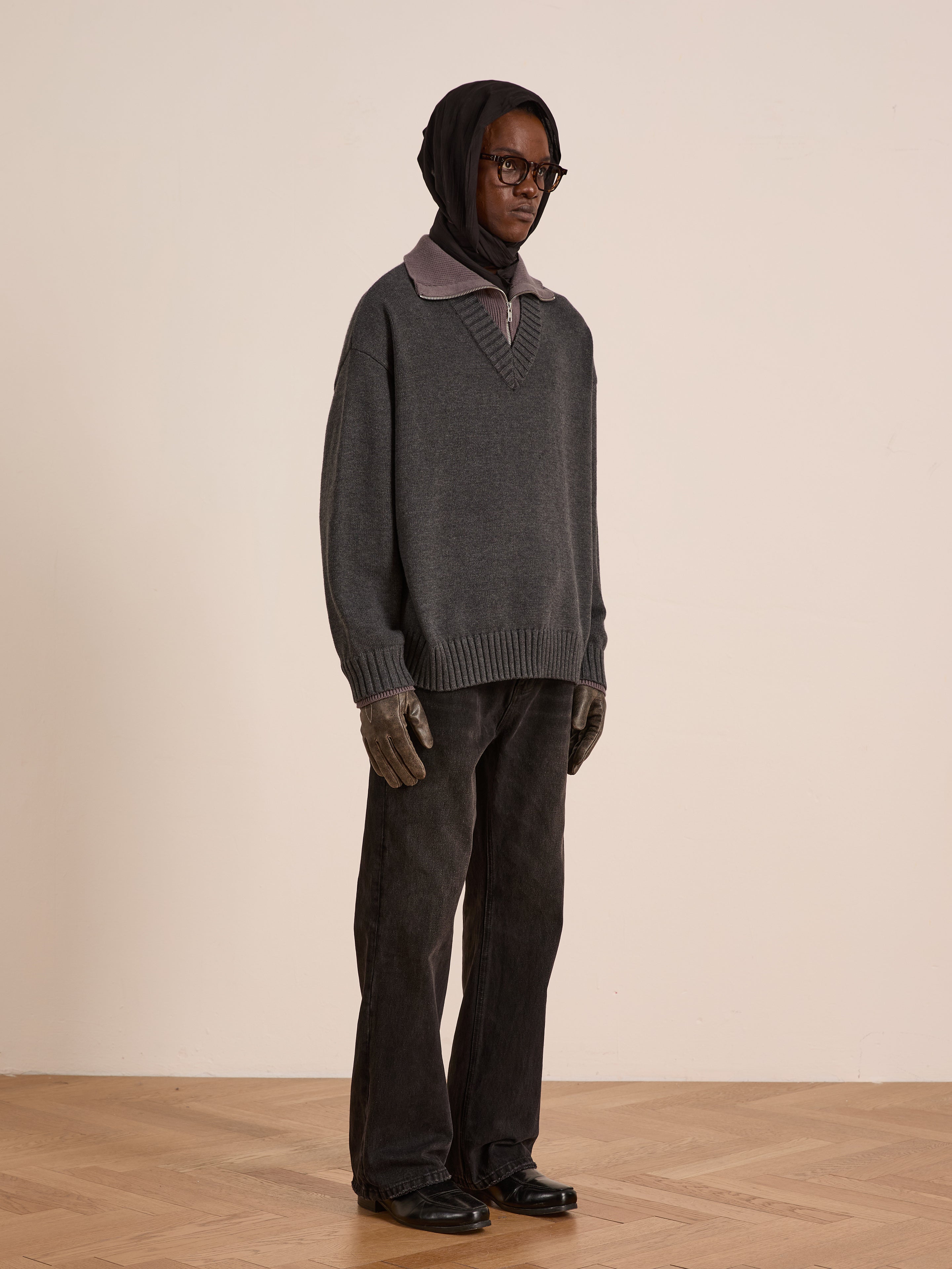 A person stands indoors on a wooden floor wearing the FOUND Double Layer Sweater—ideal for cold-weather layering—with a dark headscarf, glasses, grey wool blend gloves, collared shirt, loose dark pants, and black shoes.